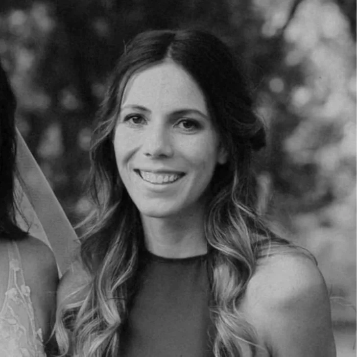 A smiling woman with long wavy hair, wearing a sleeveless dress, standing outdoors with blurred trees in the background, holding a bouquet of flowers.