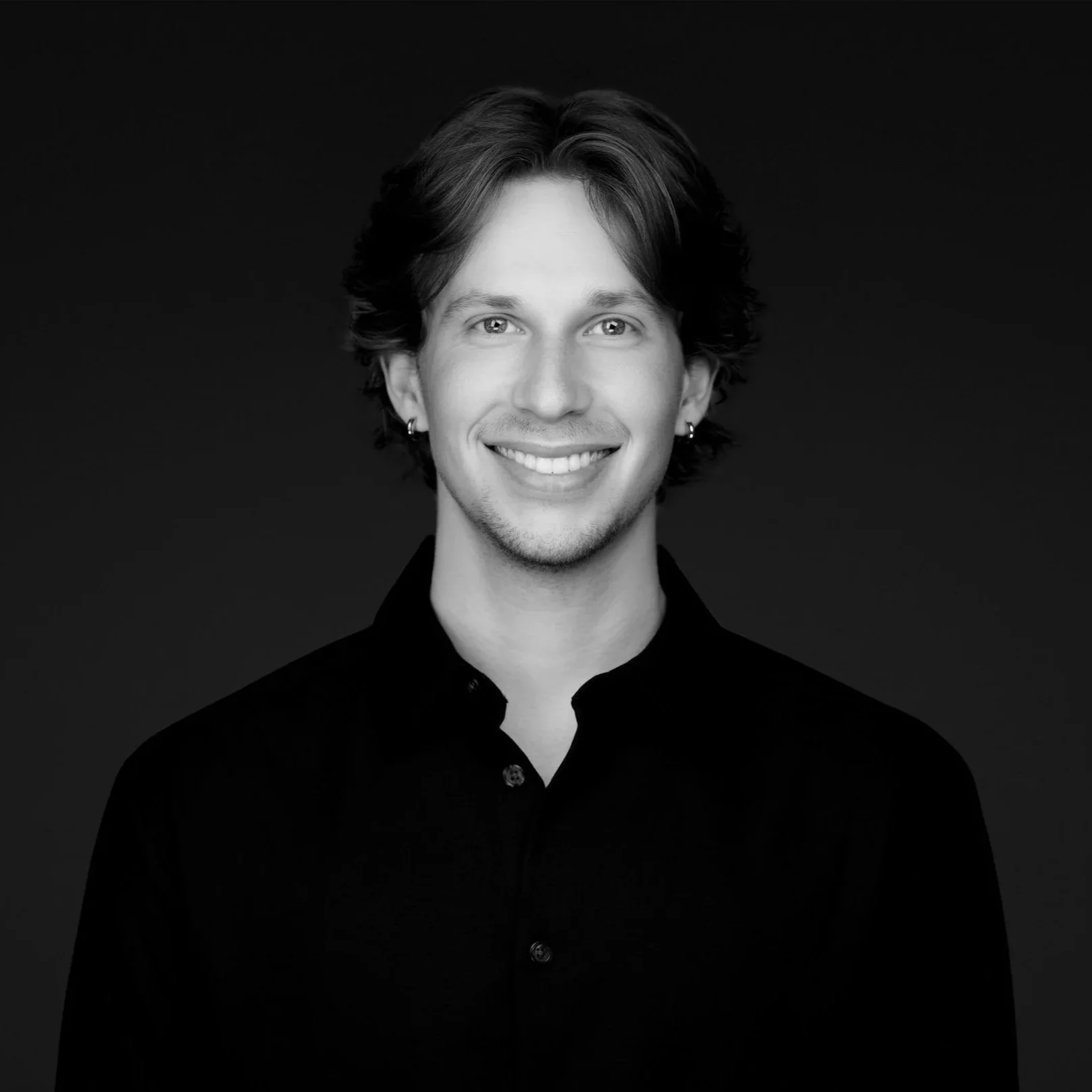 A young man with light skin, curly brown hair, blue eyes, and a mustache, smiling while wearing a gray button-up shirt and jewelry, standing against a plain white background.