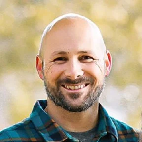 A smiling man with a beard and bald head outdoors with a blurred natural background.