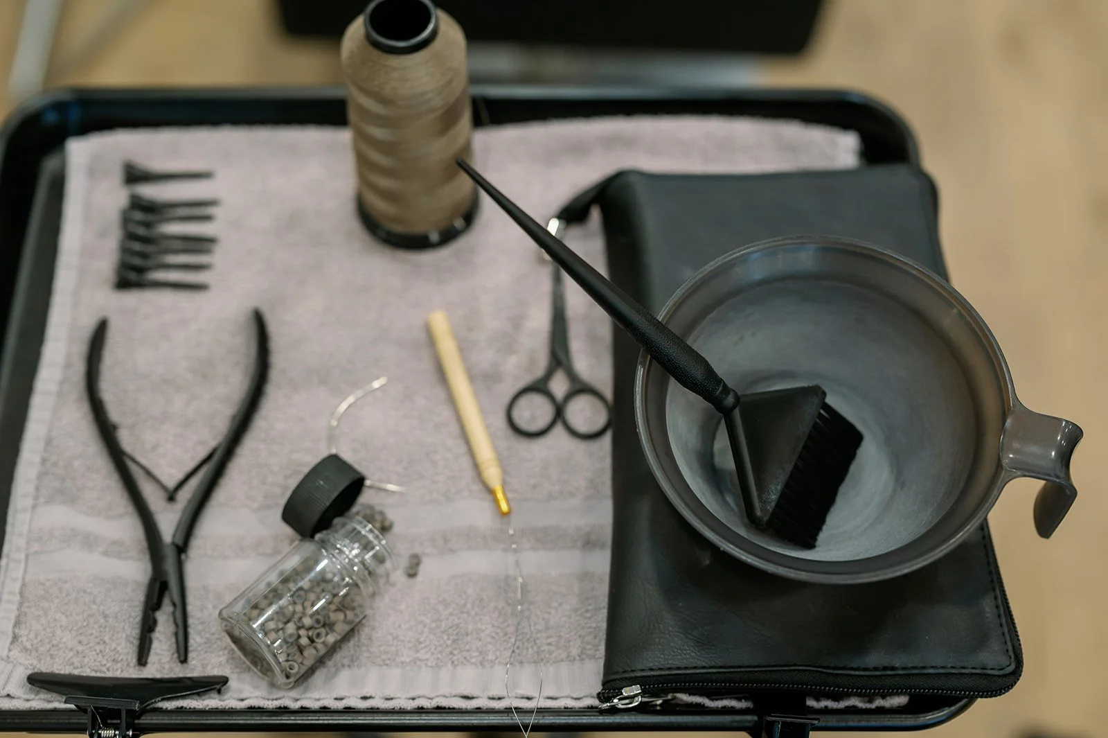 Hairdressing tools including scissors, comb, tweezers, spray bottle, threading needle, spool of thread, and a container with small hair clip beads on a towel with a bowl and brush inside.