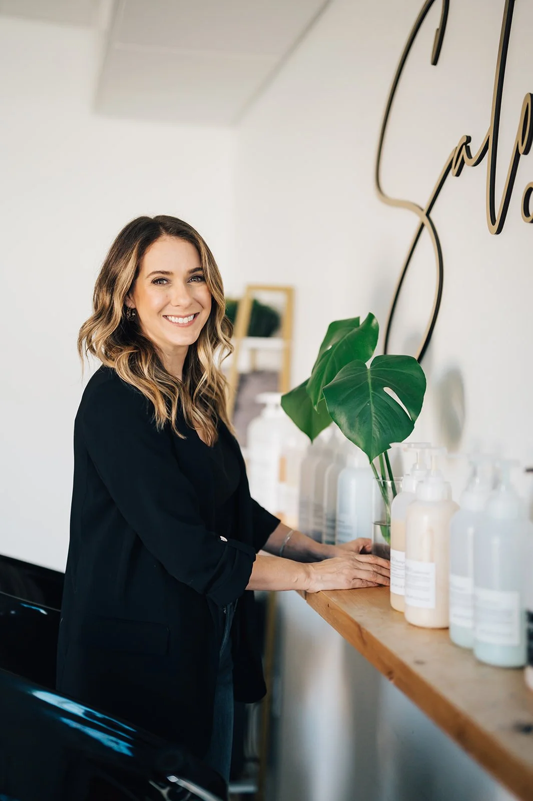 A woman with shoulder-length wavy hair smiling while standing next to a wooden shelf with bottles and a green monstera plant. She is wearing a black blazer and has a cheerful expression.
