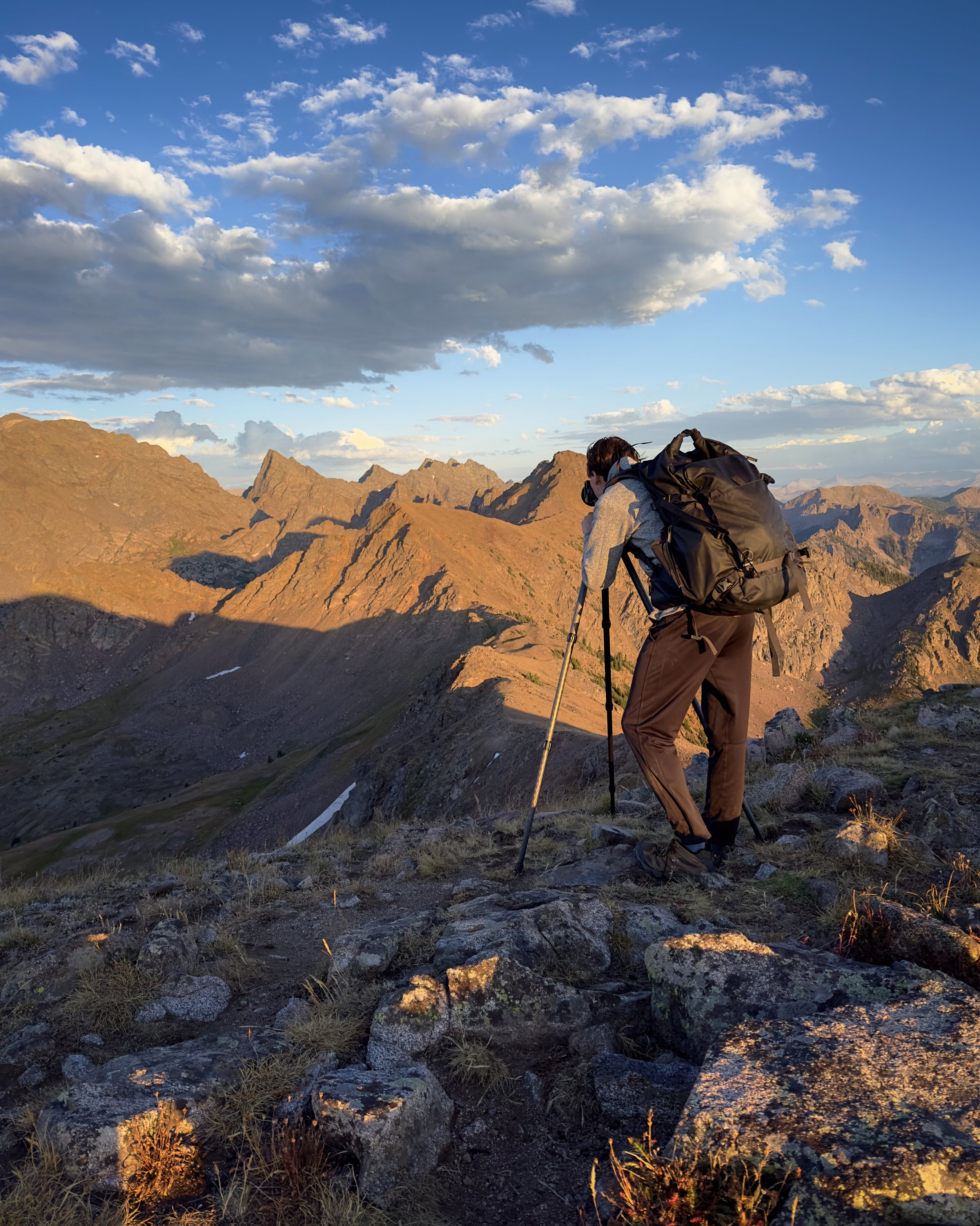 A person wearing outdoor clothing and a backpack is setting up a camera on a tripod on rocky terrain in a mountainous area. The landscape features rugged mountains under a sky with scattered clouds during sunset or sunrise.