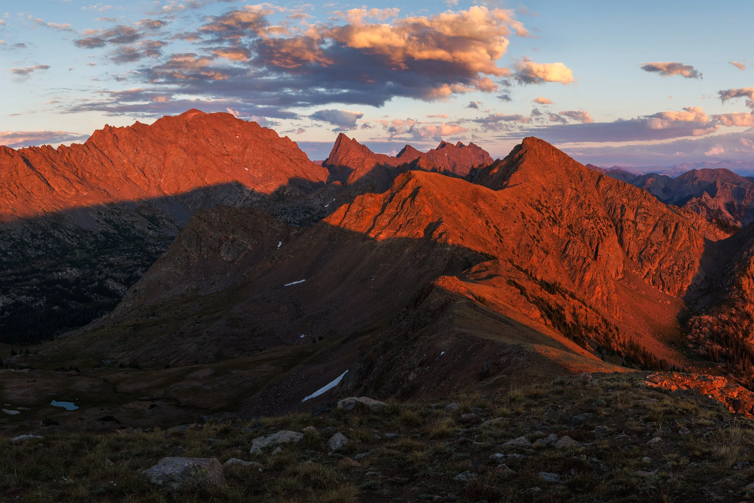 Sunset over a rugged mountain range with peaks illuminated in warm orange and red tones, under a partly cloudy sky.