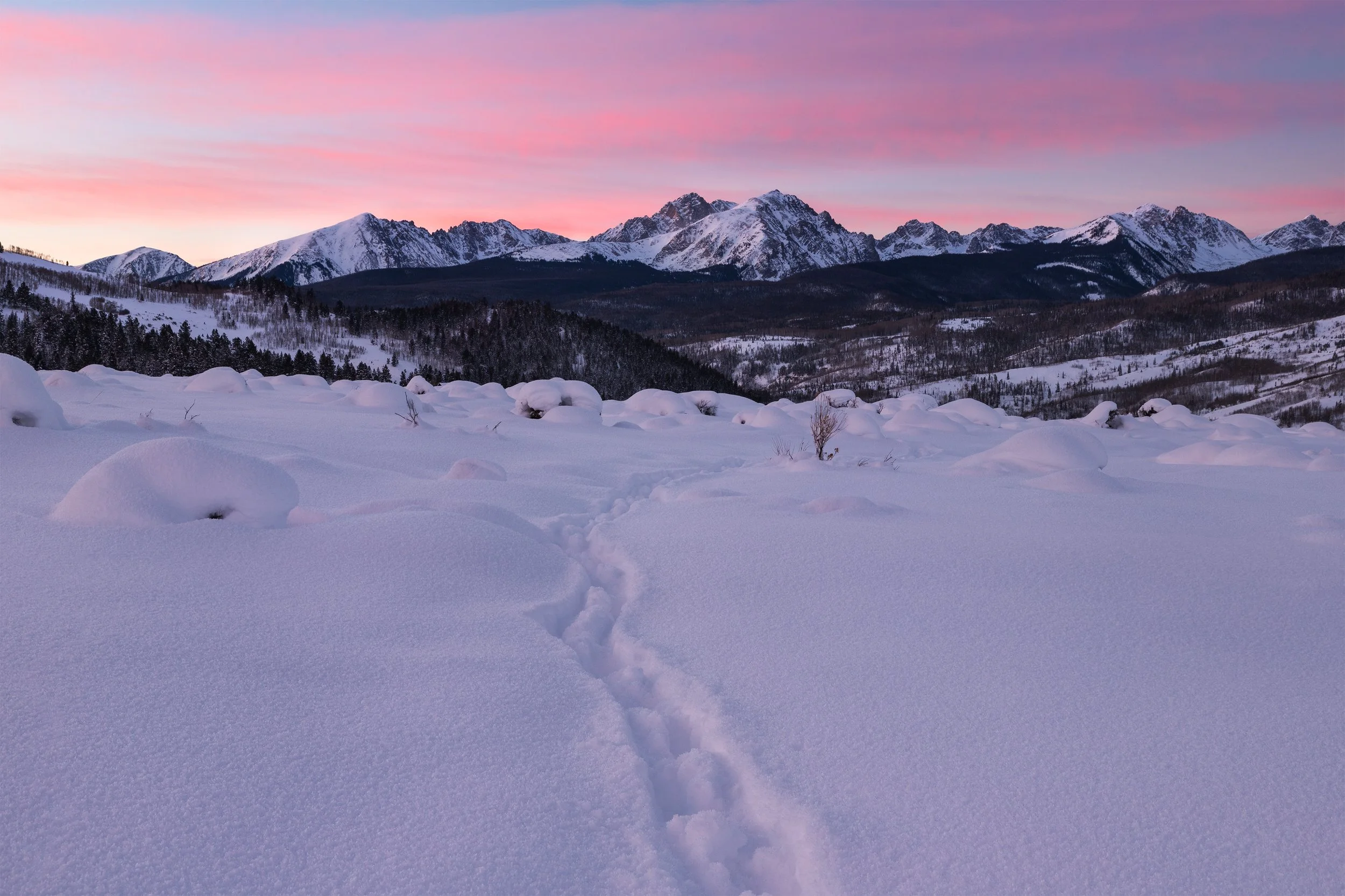 Snow-covered landscape with mountain range in the background and a pink and purple sunset sky.
