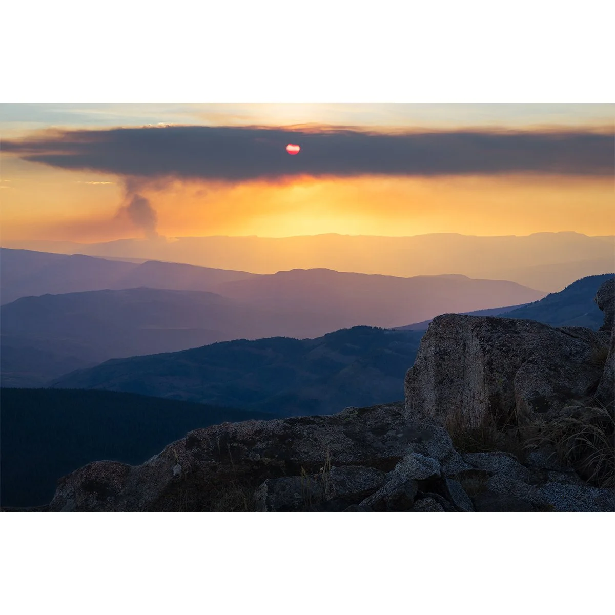 Fiery red sun setting through wildfire smoke over the distant mountains of western Colorado, viewed from the summit of Meridian Peak in the Gore Range with rocky foreground ridges.