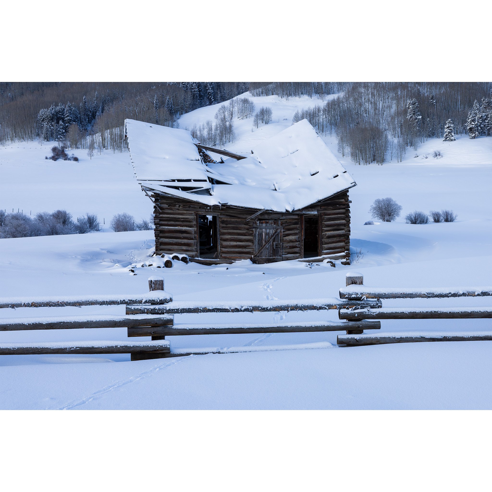 Snow-covered log cabin with a collapsed roof behind a buck-and-rail fence near Crested Butte, Colorado, in winter.
