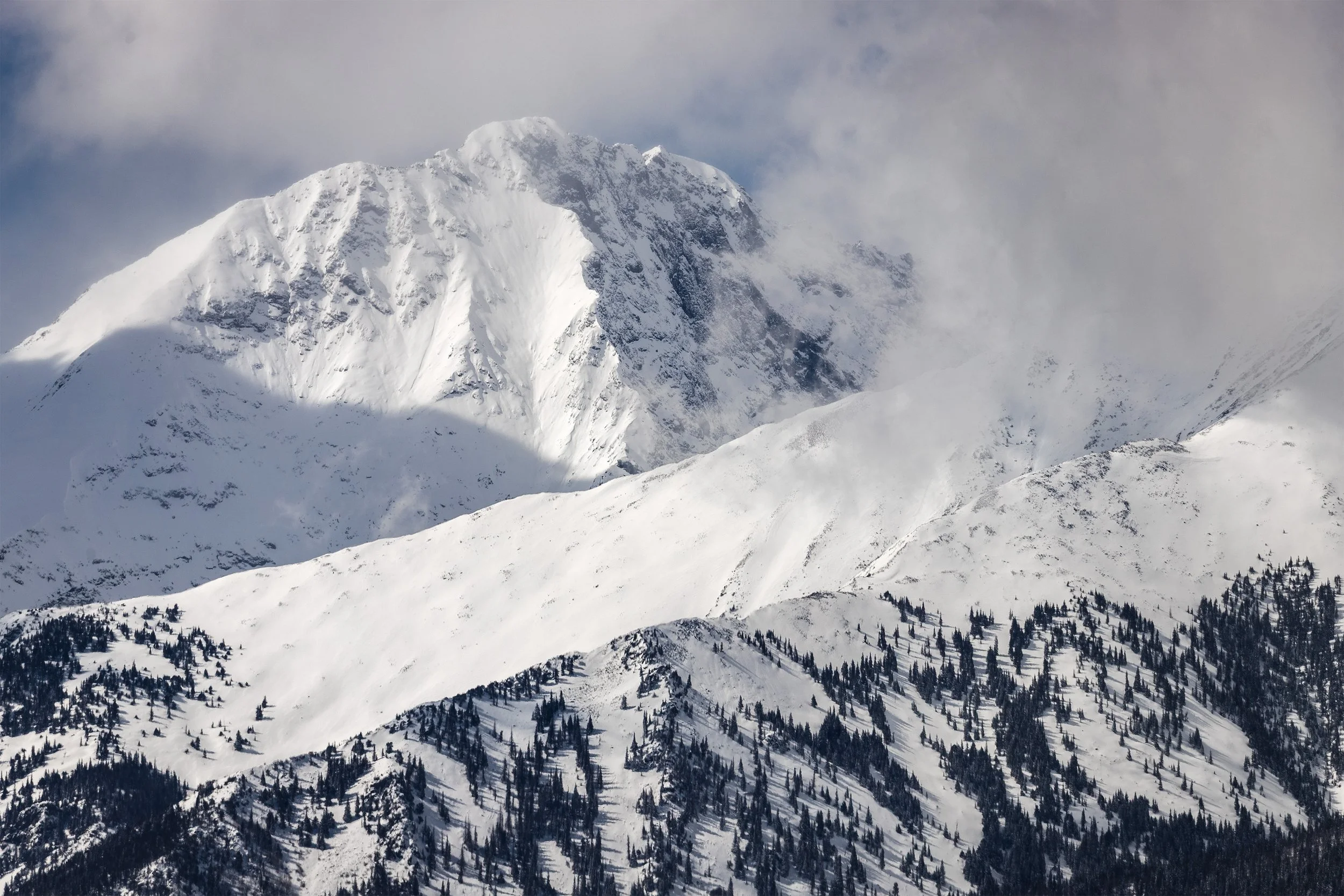 Snow-covered mountain with rugged peaks partially obscured by clouds, and a forested slope at the base.