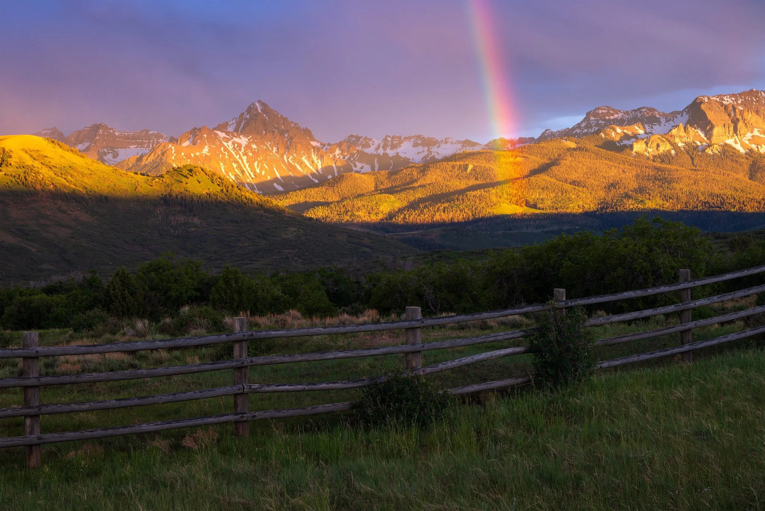 Scenic landscape of mountains with snow patches, green hills, a rainbow, and a partly cloudy sky, viewed from a grassy field with a wooden fence.