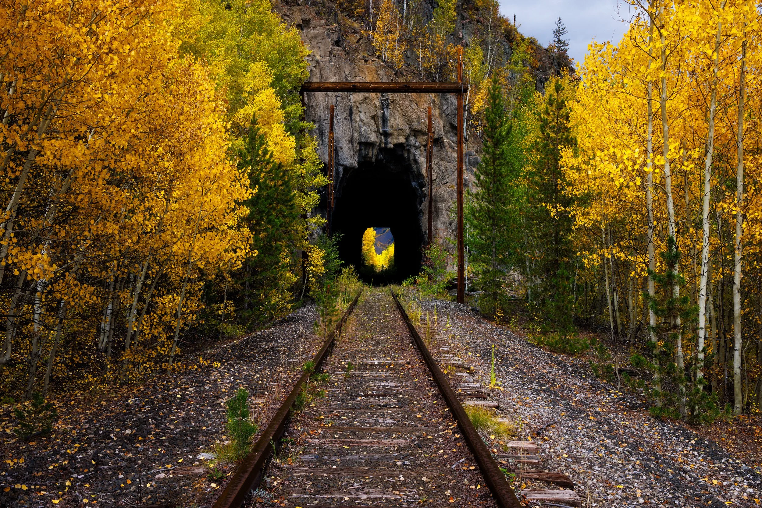 Old train tracks leading into a dark tunnel through a rocky mountain, surrounded by autumn trees with yellow and green leaves.