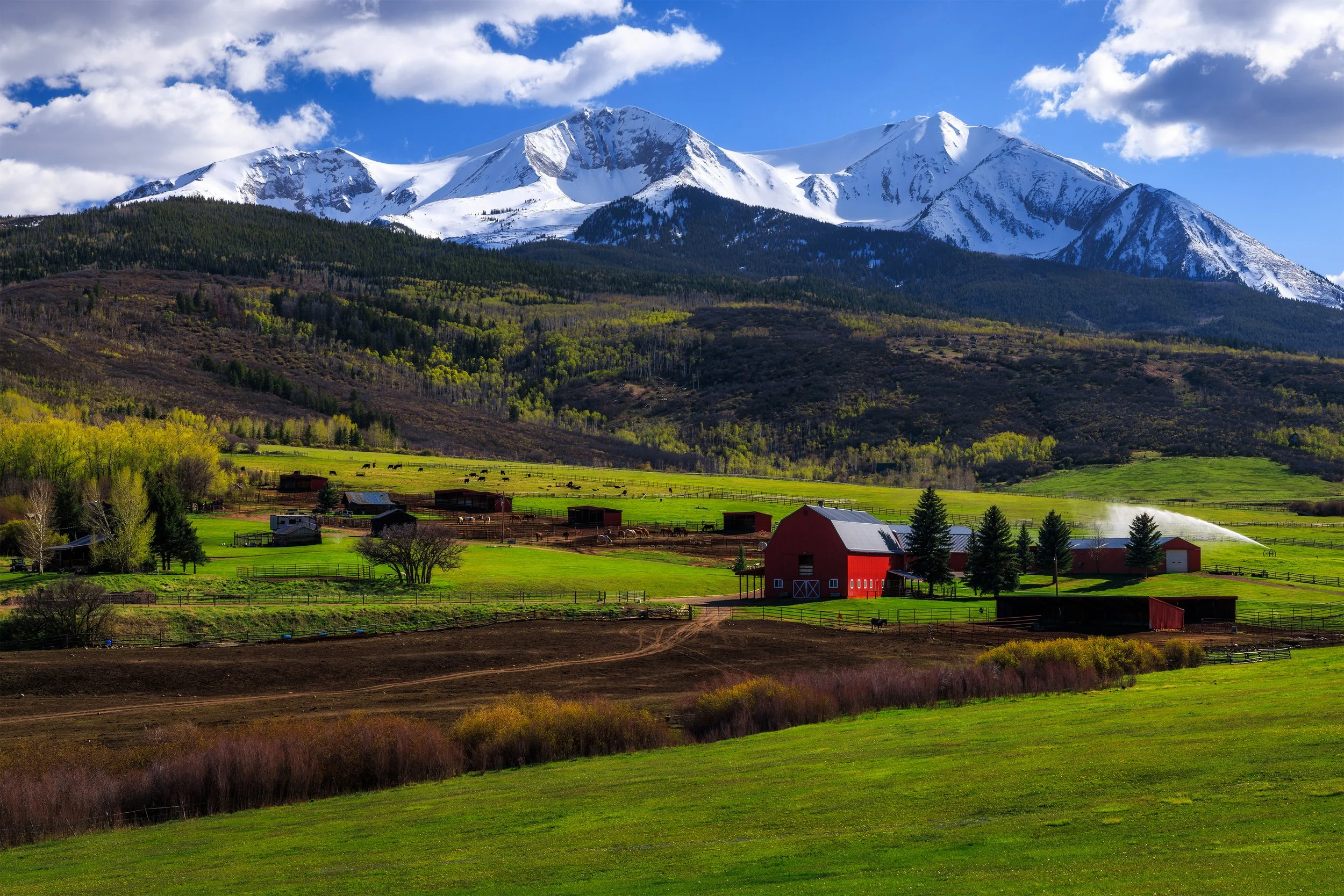 A mountain landscape with snow-capped peaks, green forested slopes, and farm buildings in the foreground, including a red barn and green fields with grazing animals, under a partly cloudy blue sky.