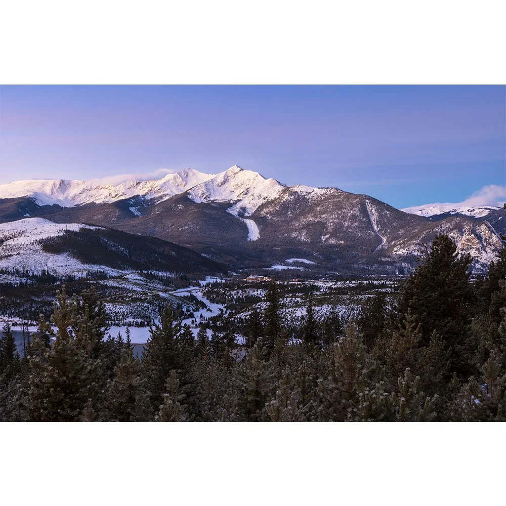 The snow-covered Tenmile Range near Frisco, Colorado glows in soft pre-dawn light above a wintery valley with distant town lights and forested hills