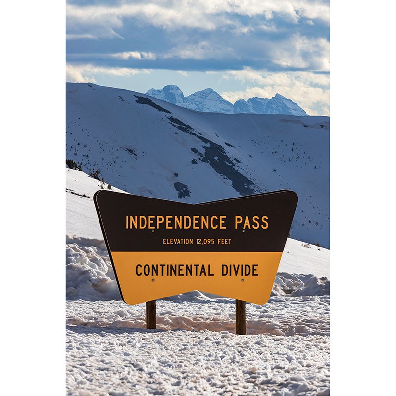 A snow-covered Independence Pass sign at 12,095 feet with the Maroon Bells and Pyramid Peak visible in the distance under a partly cloudy sky.