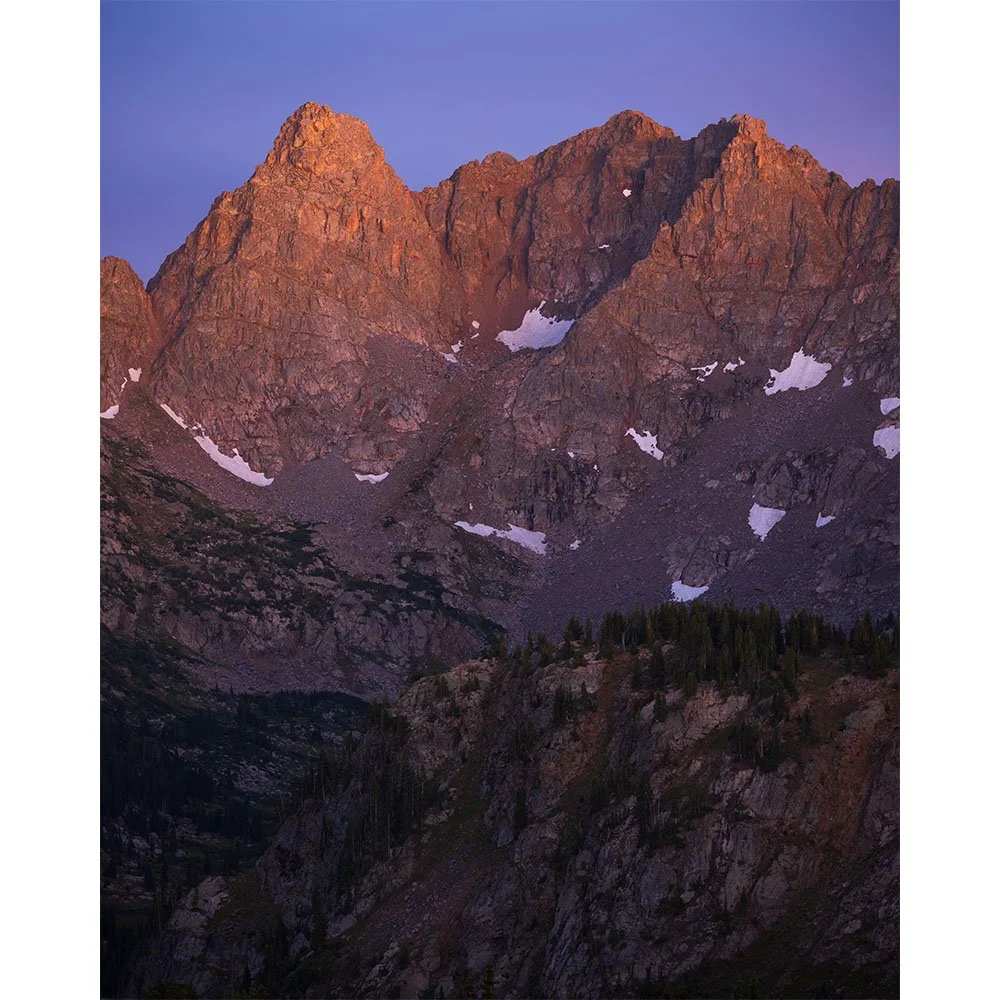 Rugged unnamed peaks in the Gore Range of Colorado glowing with alpenglow after sunset, with steep granite cliffs, snow patches, and a deep alpine valley below.