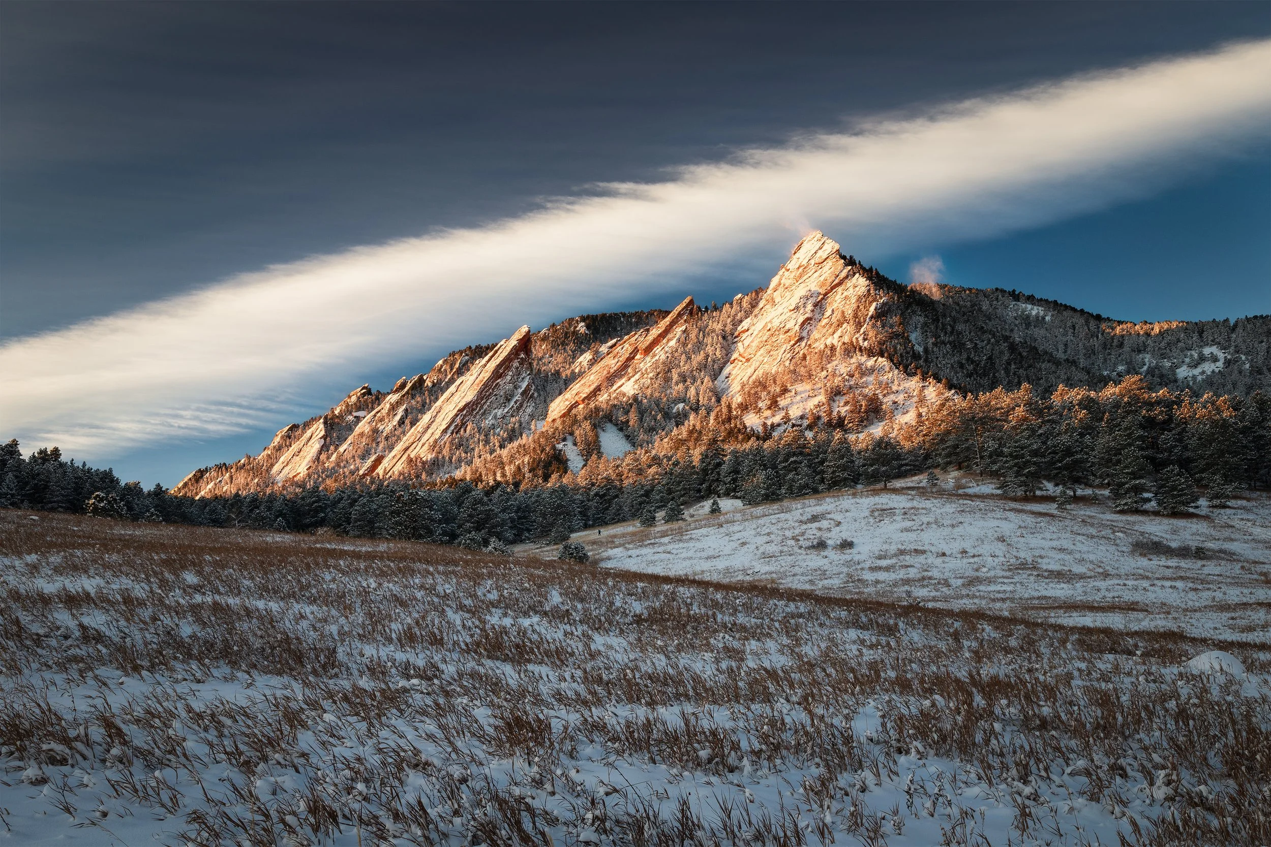 Snow-covered field with tall brown grass in the foreground, forested hillside, and jagged mountain peaks illuminated by sunlight under a partly cloudy sky.