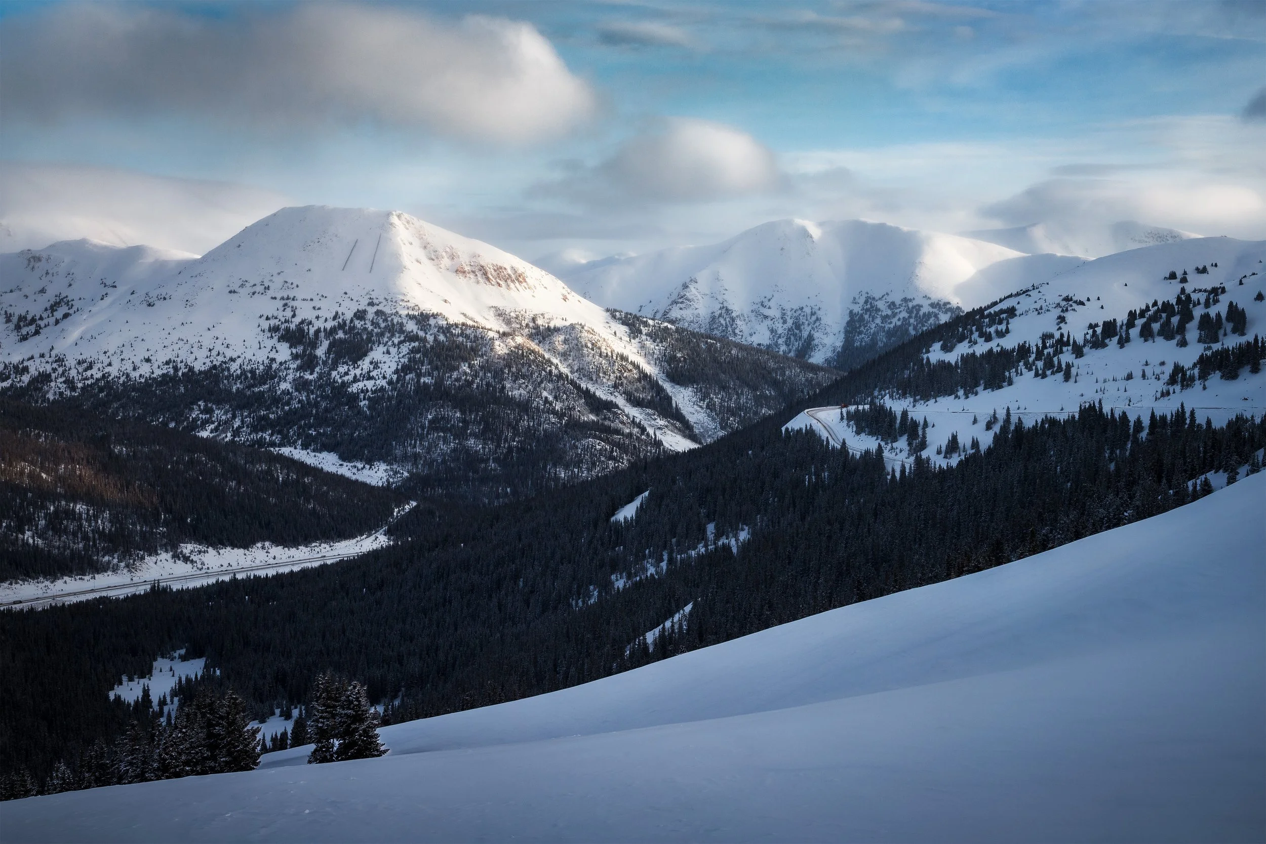Snow-covered mountains with a mix of evergreen trees and open snowy slopes under a partly cloudy sky.