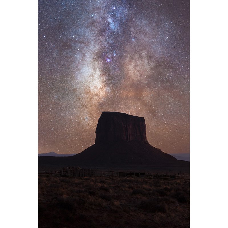 The Milky Way rises behind a large sandstone butte in Monument Valley, Arizona, under a starry sky filled with galactic detail.