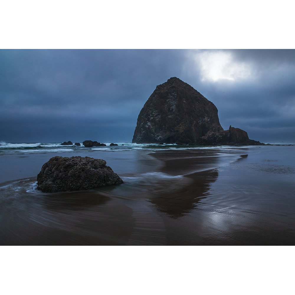 A moody, overcast evening at Cannon Beach on the Oregon Coast, featuring the iconic Haystack Rock rising from the ocean with gentle waves swirling around foreground rocks on the wet sand.