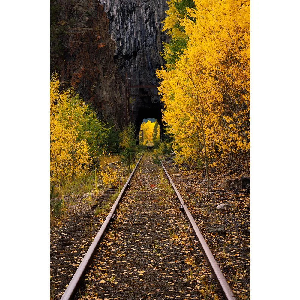Abandoned railroad tracks leading into a mountain tunnel surrounded by bright yellow aspen trees in full autumn color near Eagle County, Colorado.
