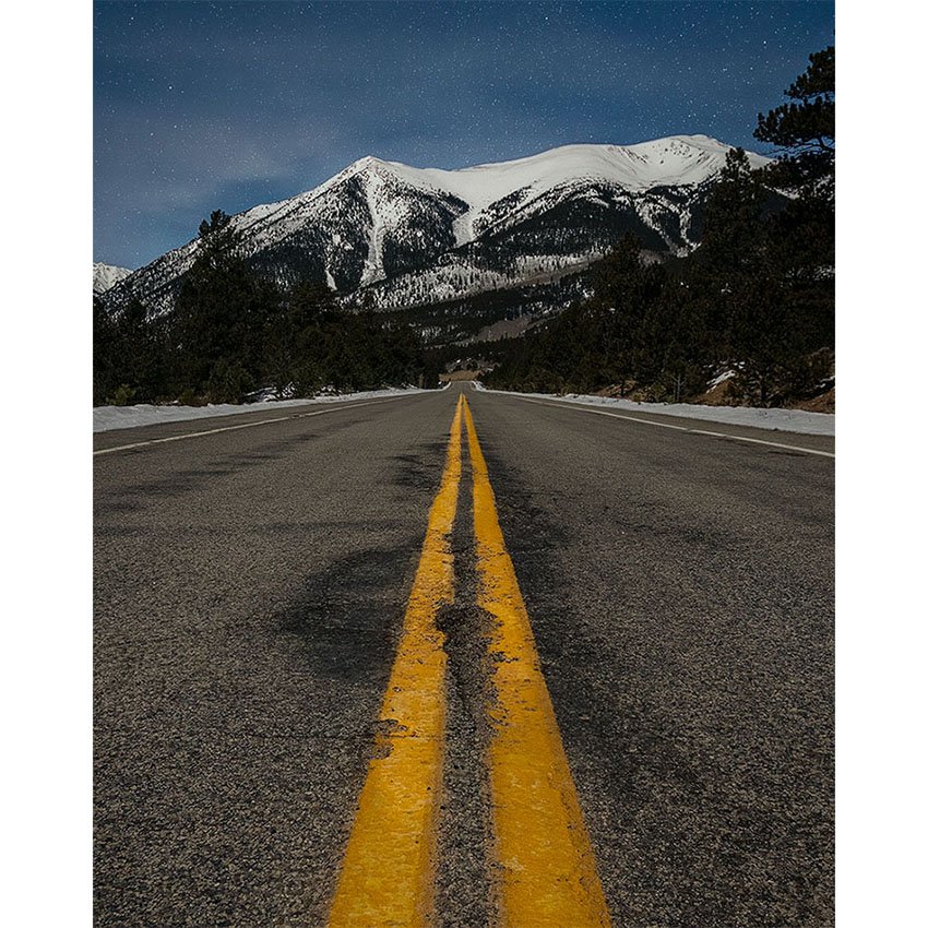 View down a highway centerline toward snow-covered Mount Elbert under a starry night sky near Twin Lakes, Colorado.