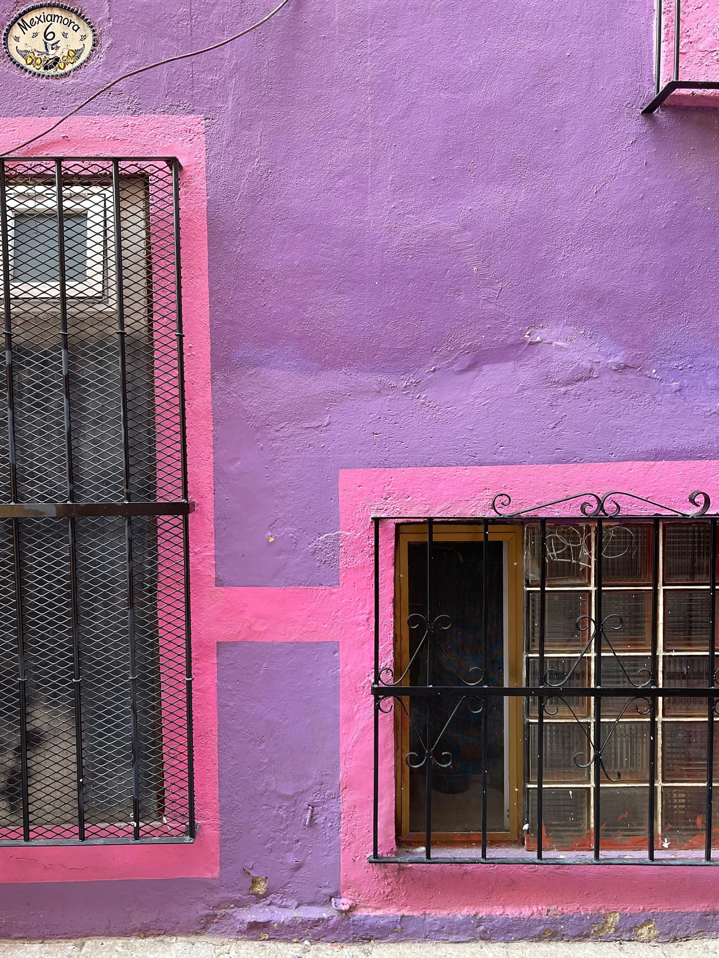 Colorful wall with a purple and pink paint scheme, featuring two barred windows. A decorative sign reads "Mexiamora 6" above the left window.