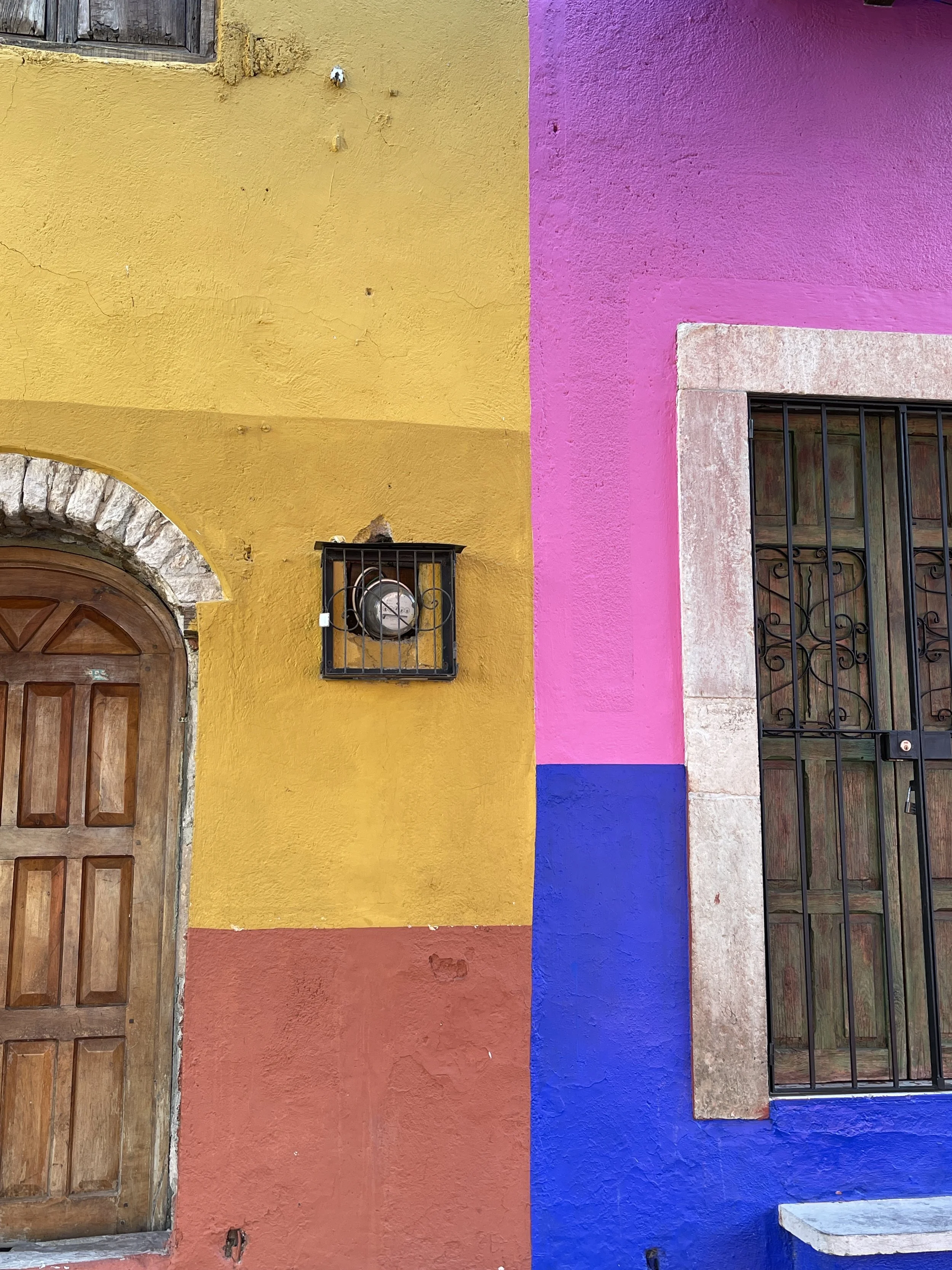 Colorful stucco wall with yellow, pink, red, and blue sections, featuring a wooden door with an arched stone frame on the left and a metal-barred window on the right.