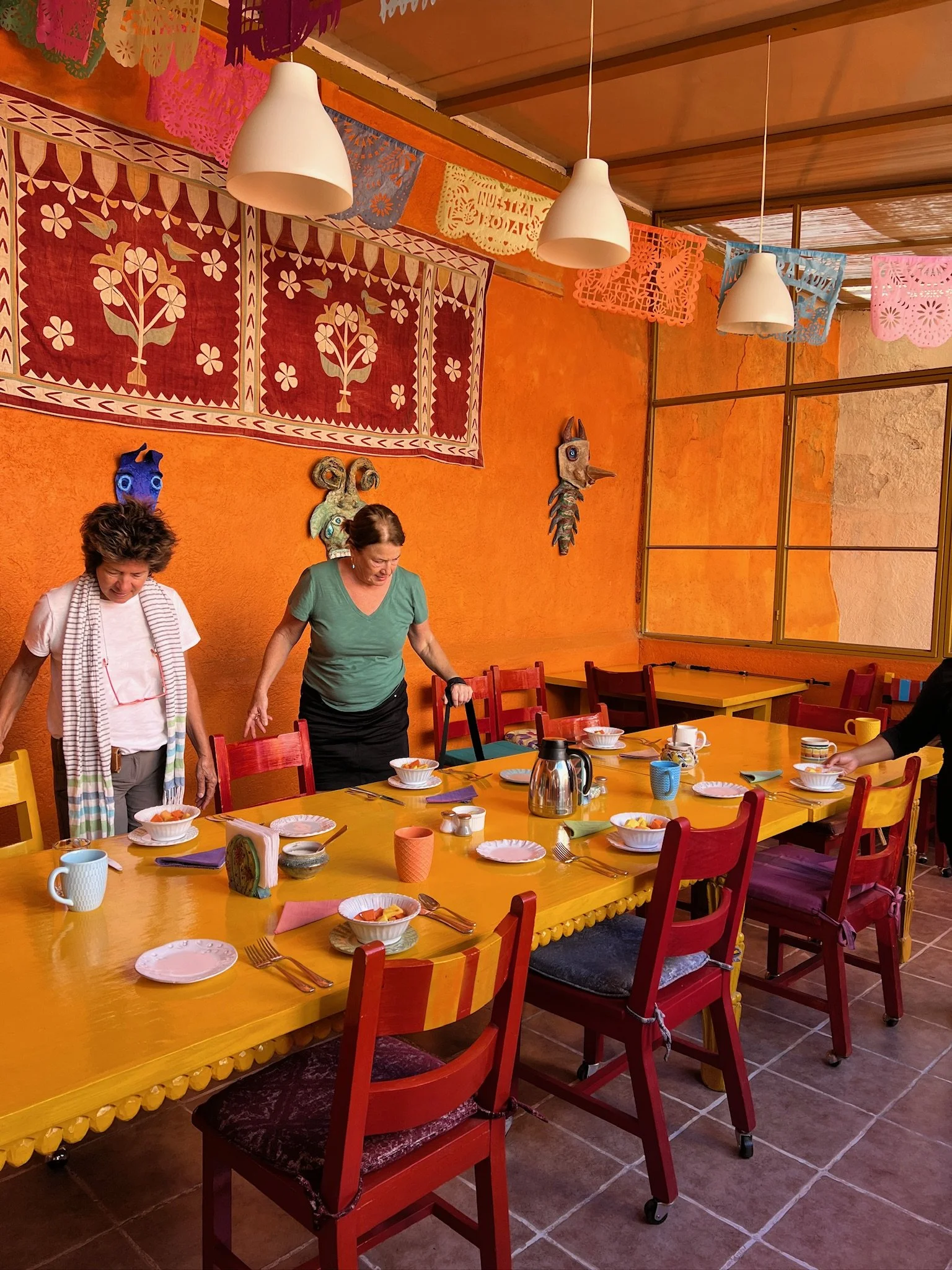 Colorful dining room with orange walls, decorated with Mexican papel picado banners, a large yellow table set with plates and mugs, and two people setting the table.