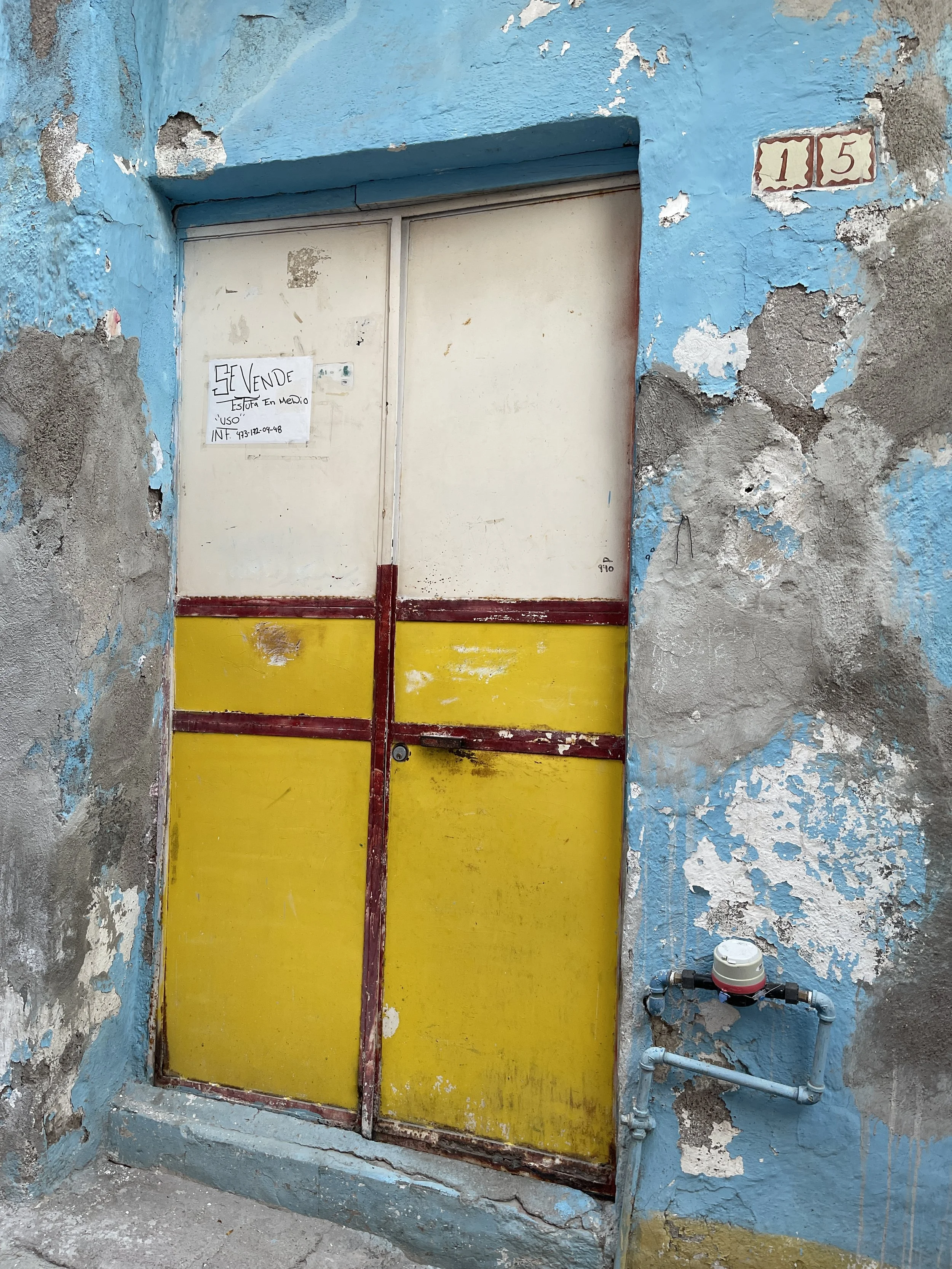 Old, weathered blue wall with a deteriorating door painted in yellow and white, featuring a handwritten sale notice. The wall has peeling paint and displays number 15. There's a water meter attached nearby.
