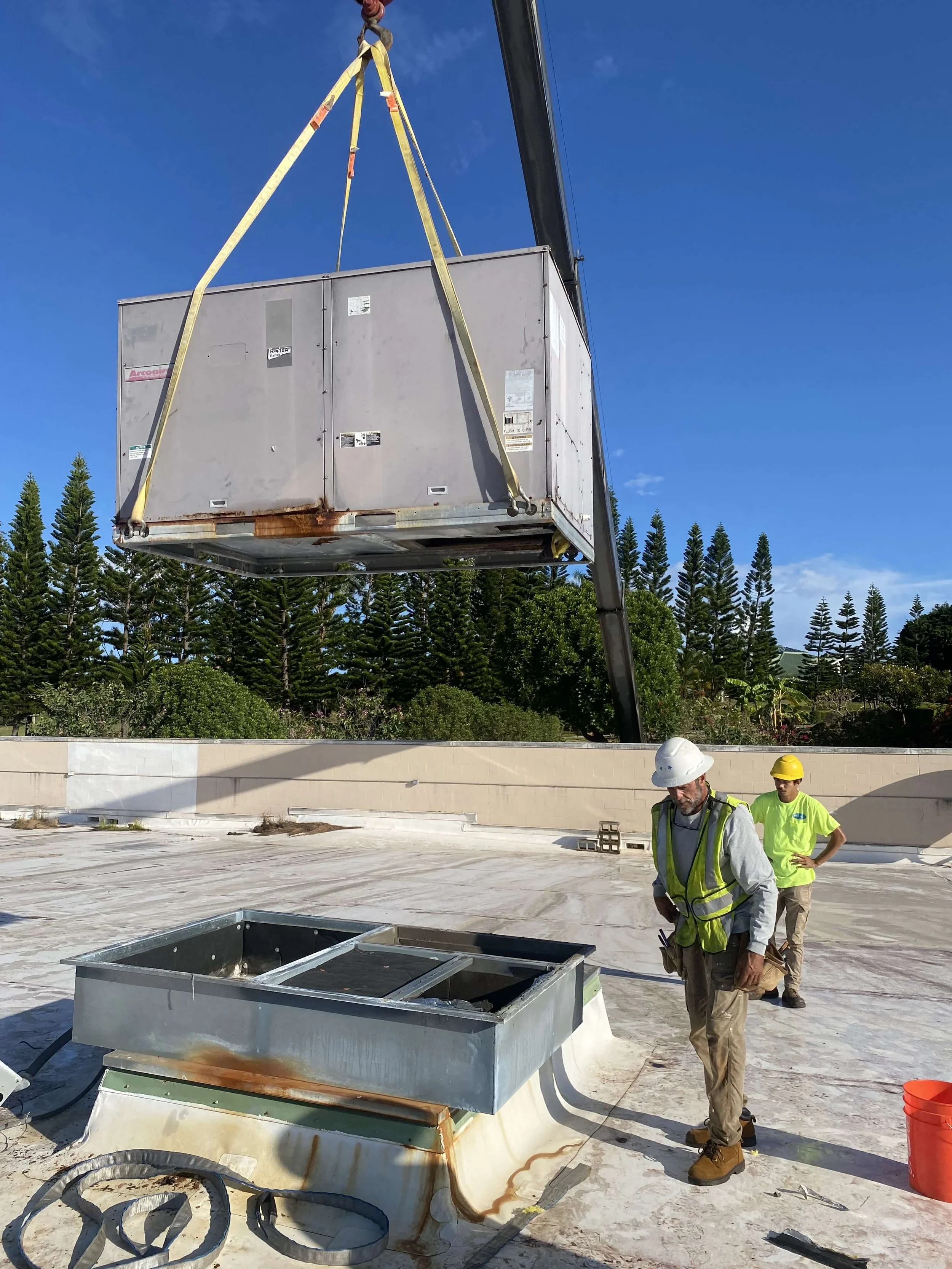 An old HVAC unit is suspended high above the rooftop by a crane during the removal operation