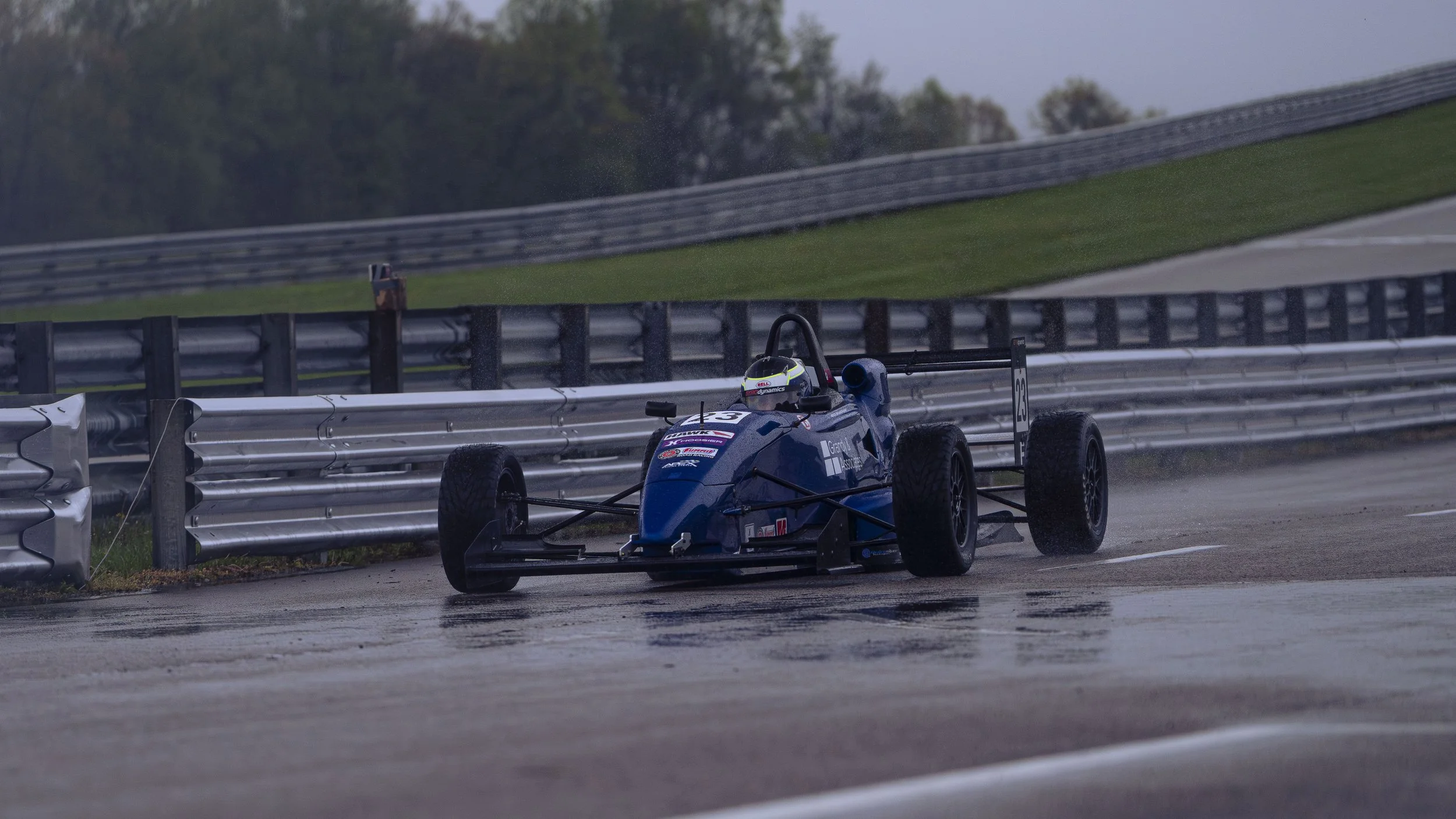 A blue single-seat race car driving off a wet road near a metal guardrail, with trees and a cloudy sky in the background.
