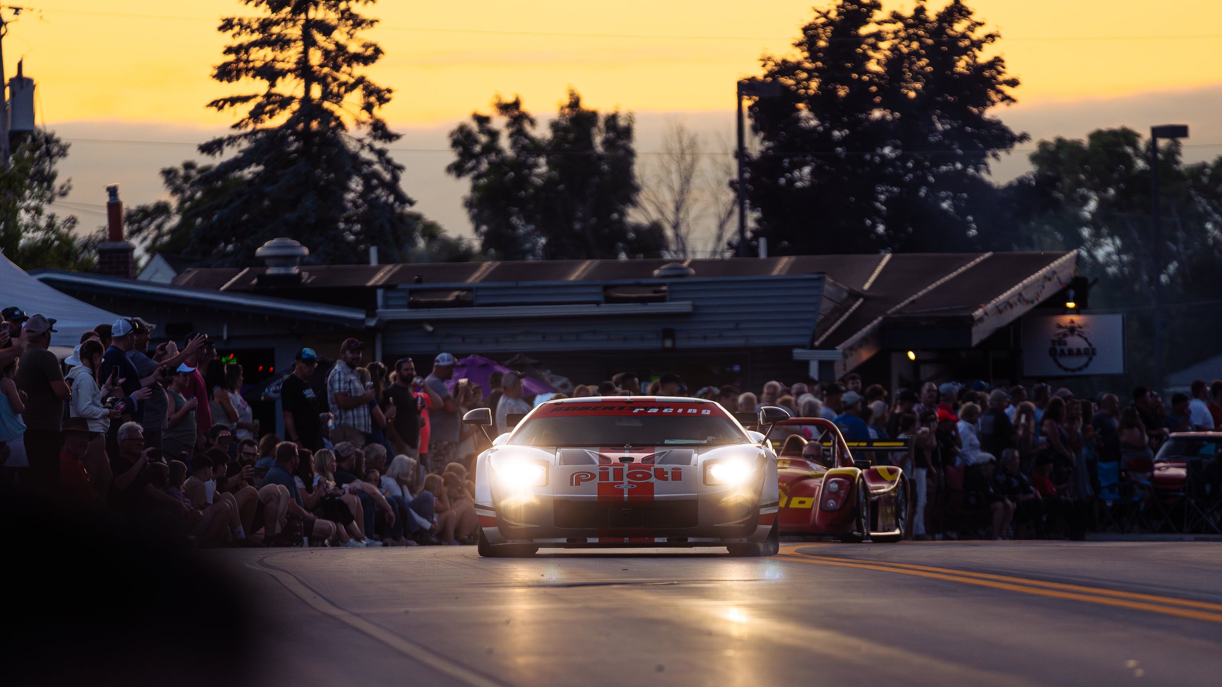 A racing car driving down a street at sunset, with a large crowd of spectators watching on both sides. The scene shows a lively motorsports event with the car emitting bright headlights and branding, such as Pirelli.