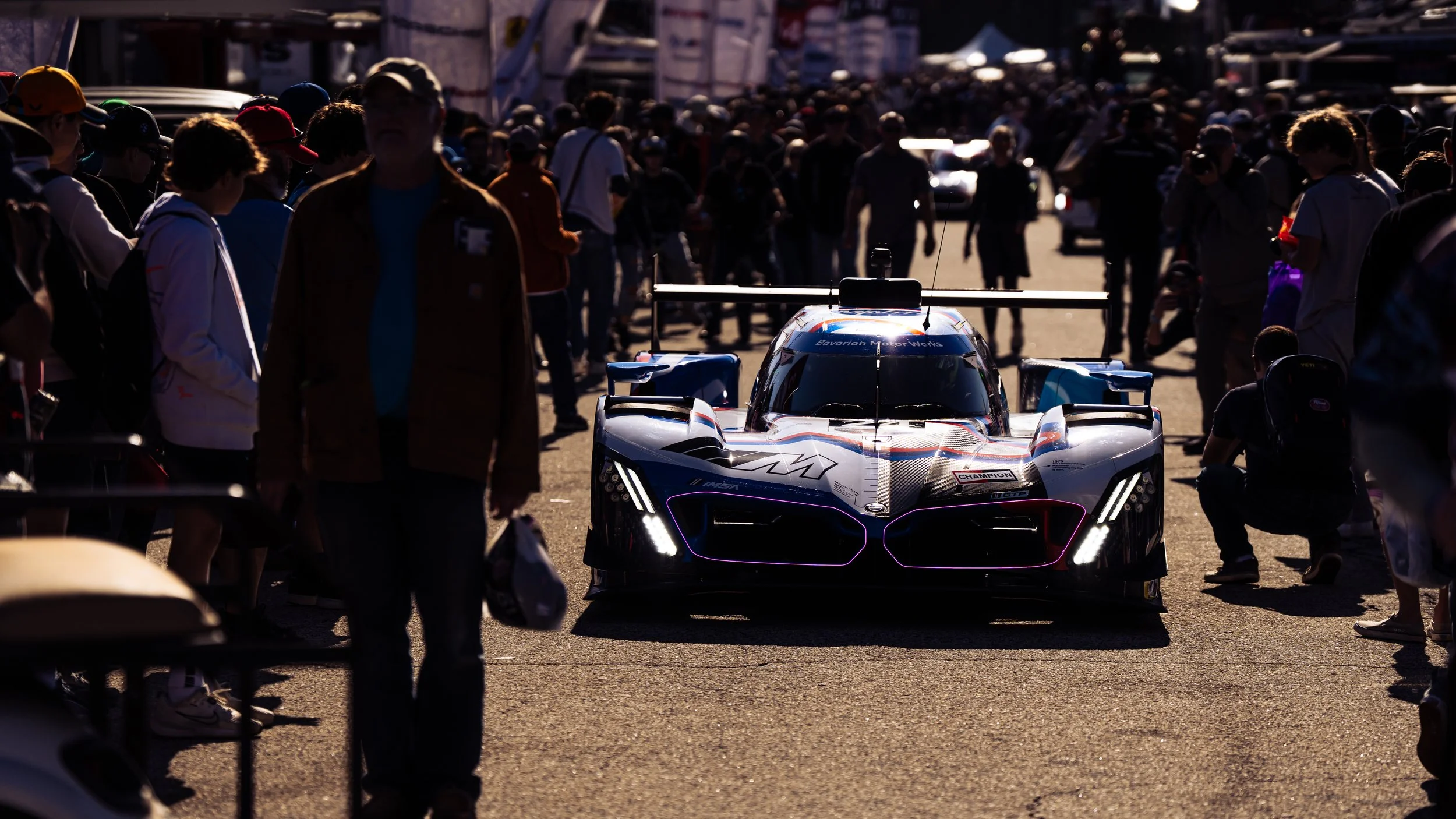 A race car with a sleek black and white design and vibrant neon accents, surrounded by a crowd of spectators in the pit lane area at a racing event.
