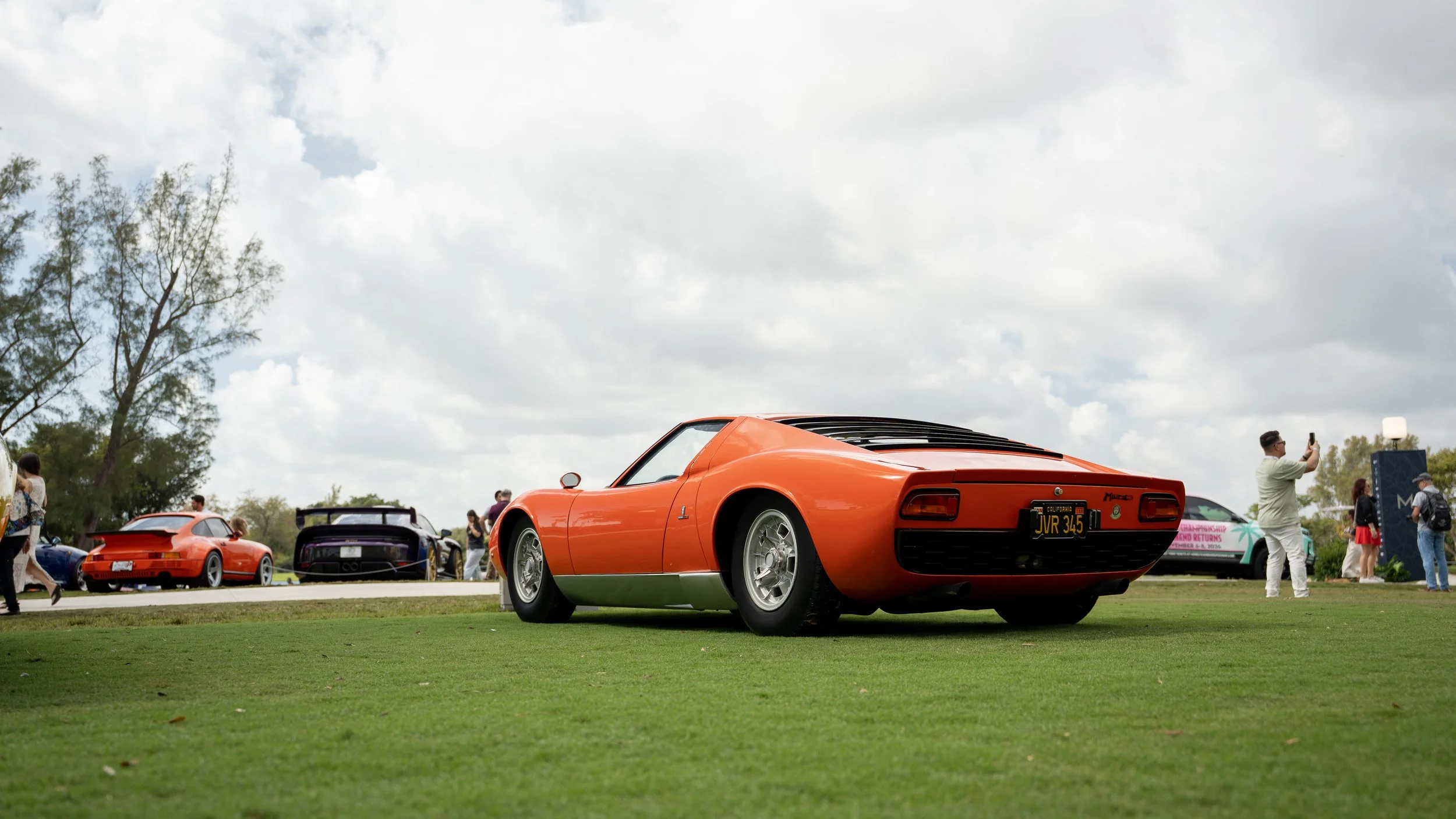 A bright orange classic sports car parked on a grass field at an outdoor car show, with a crowd of people and other cars in the background under a partly cloudy sky.