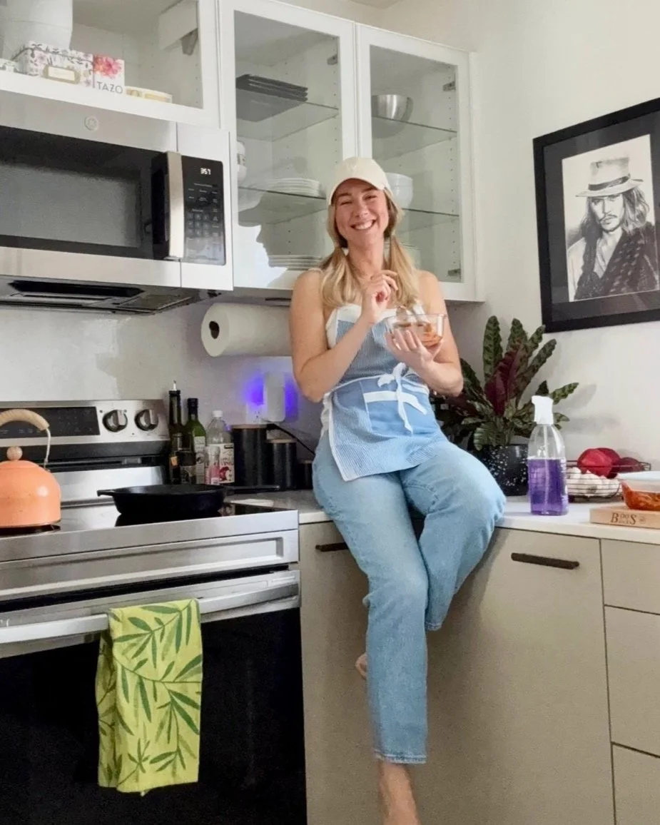 Person in a kitchen sitting on a counter, wearing a cap and apron, holding a bowl, with a stove, microwave, and plant nearby.