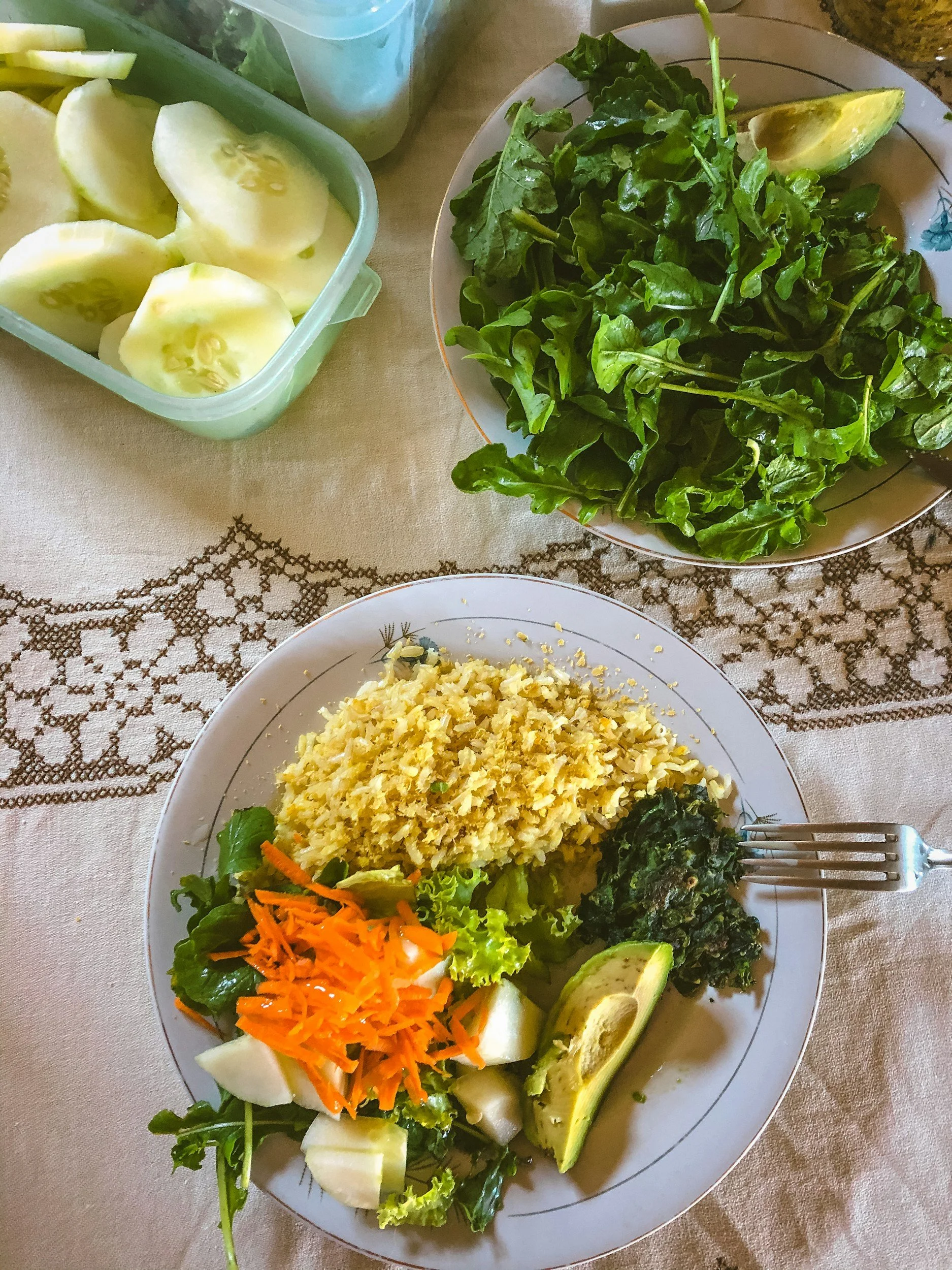 A plate of rice, mixed vegetables including carrots, and leafy greens, with avocado slices on the side. In the background, there is a bowl of chopped cucumbers and a container of sliced cucumber with seeds visible.