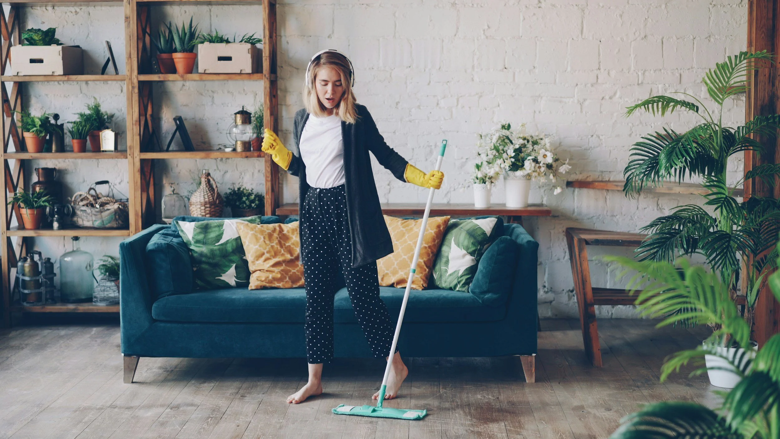 A woman cleaning an indoor space with a mop. She is wearing yellow rubber gloves, a white shirt, a dark cardigan, and black polka-dot pants. The room features a blue sofa with patterned pillows, wooden shelves with potted plants, and a white brick wall.