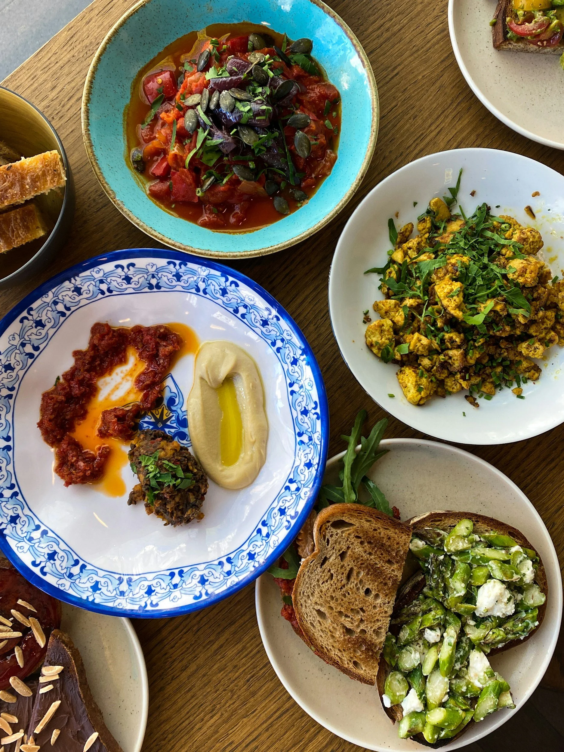 Various dishes of food on a wooden table, including a plate of scrambled eggs with herbs, a bowl of tomato and vegetable stew topped with seeds, a plate of avocado toast with cheese, and a dessert with chocolate and nuts.