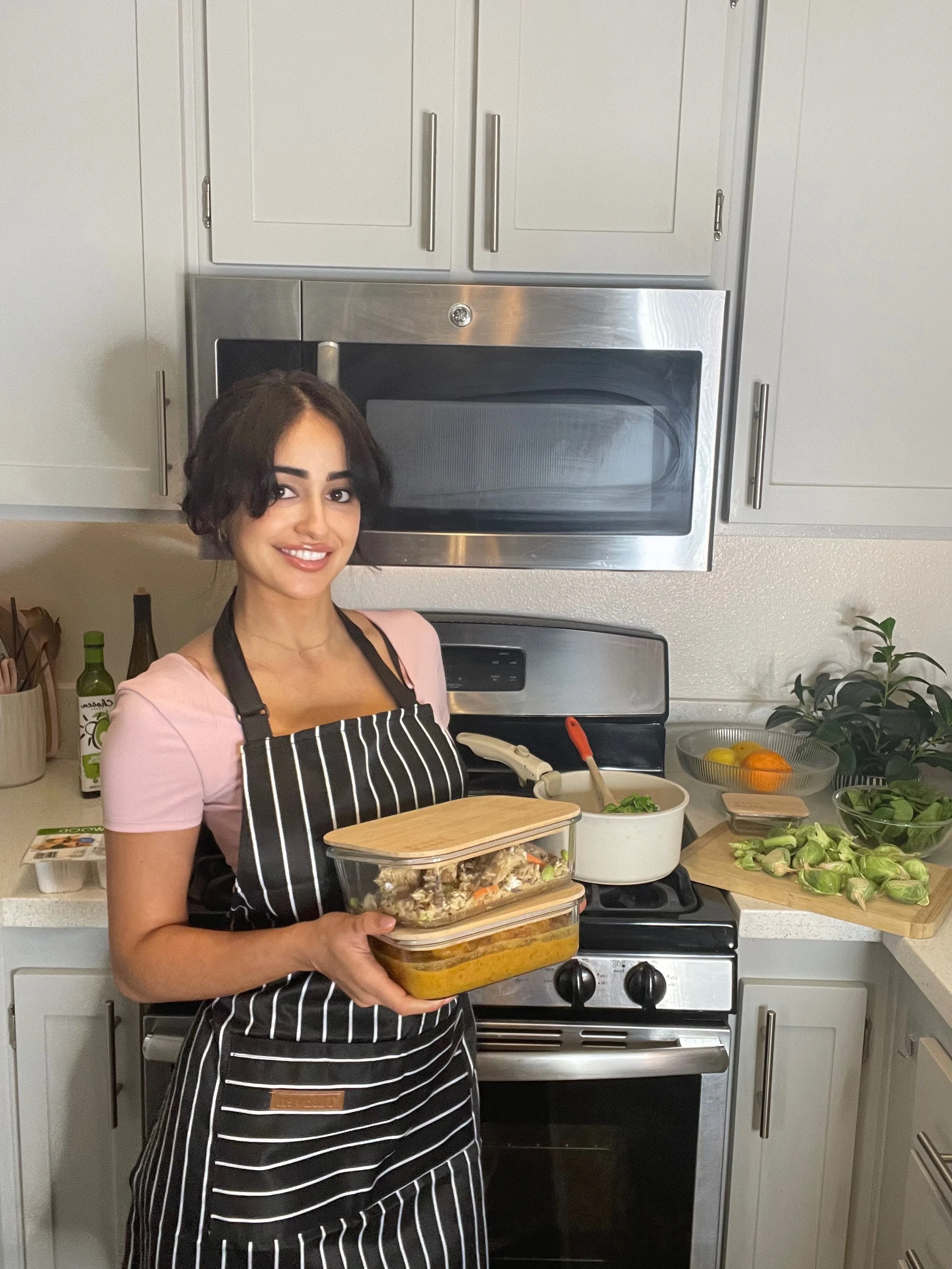 Person in a kitchen holding food containers, wearing a striped apron in front of a stove with cooking utensils and ingredients.