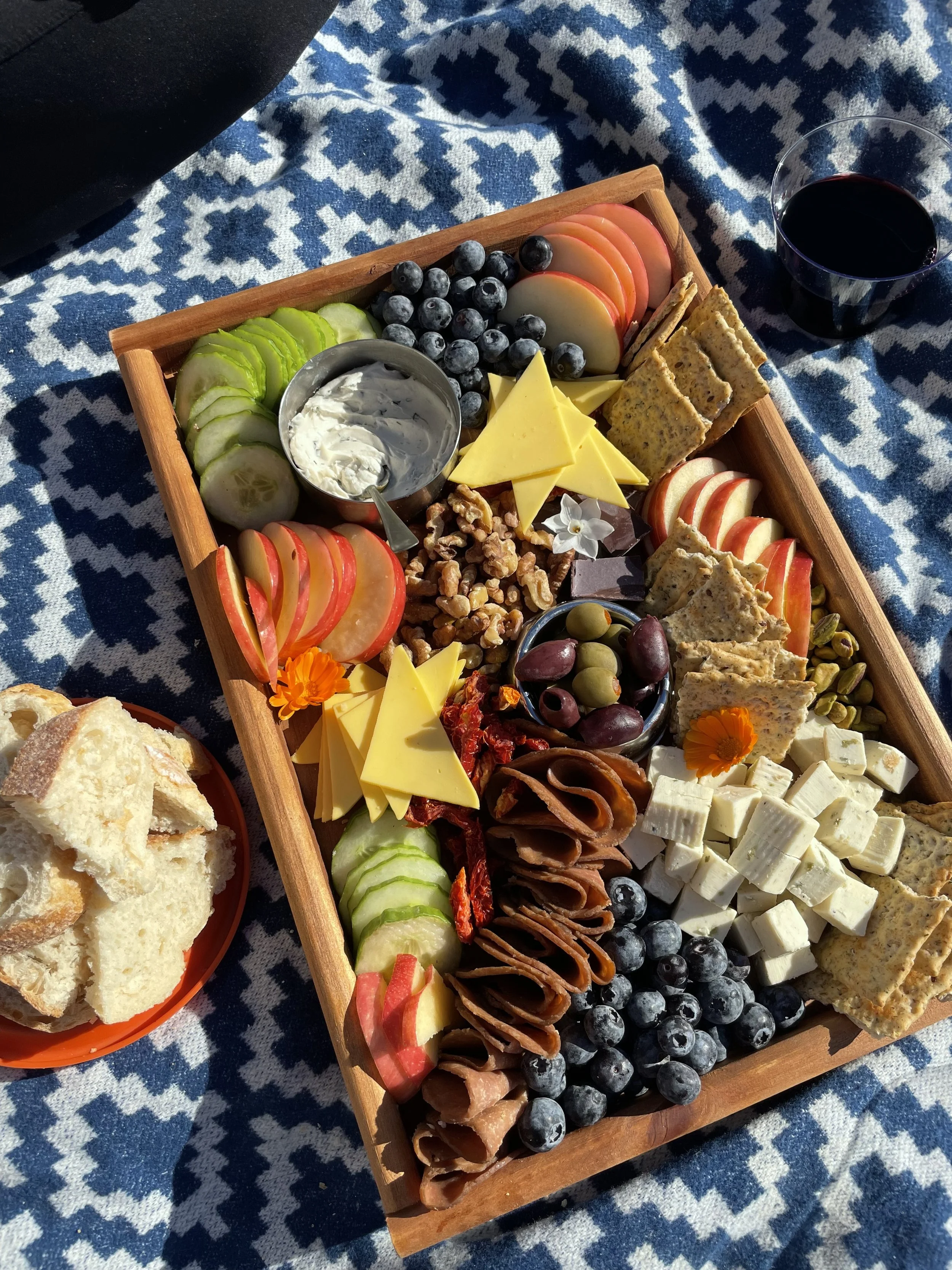 A wooden serving tray with assorted cheeses, blueberries, cucumber slices, sliced apple, crackers, walnuts, grapes, sun-dried tomatoes, and a small bowl of spread, placed on a blue and white patterned cloth next to a glass of red wine and a slice of bread.