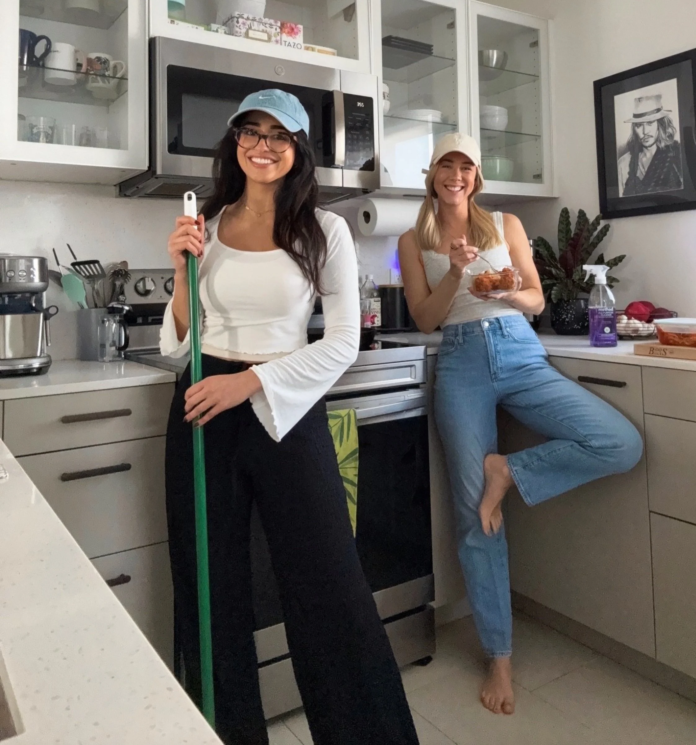 Two smiling women in a kitchen, one wearing a blue baseball cap and glasses holding a broom, and the other wearing a white cap holding a bowl of food, with kitchen cabinets, microwave, and framed artwork in the background.