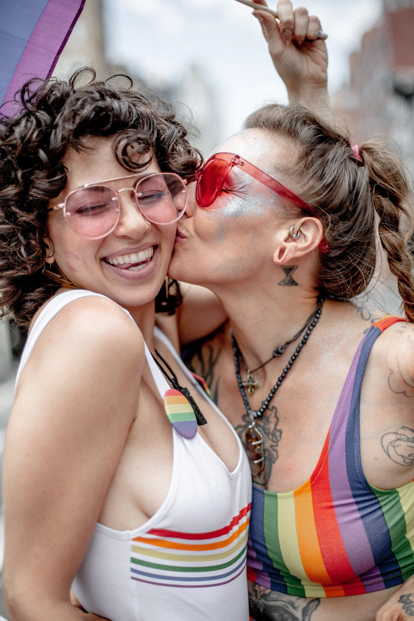 Two queer people smiling, wearing colorful rainbow-themed clothing and sunglasses, celebrating at a pride event. One person is kissing the other on the cheek after attending couples therapy for queer folk.
