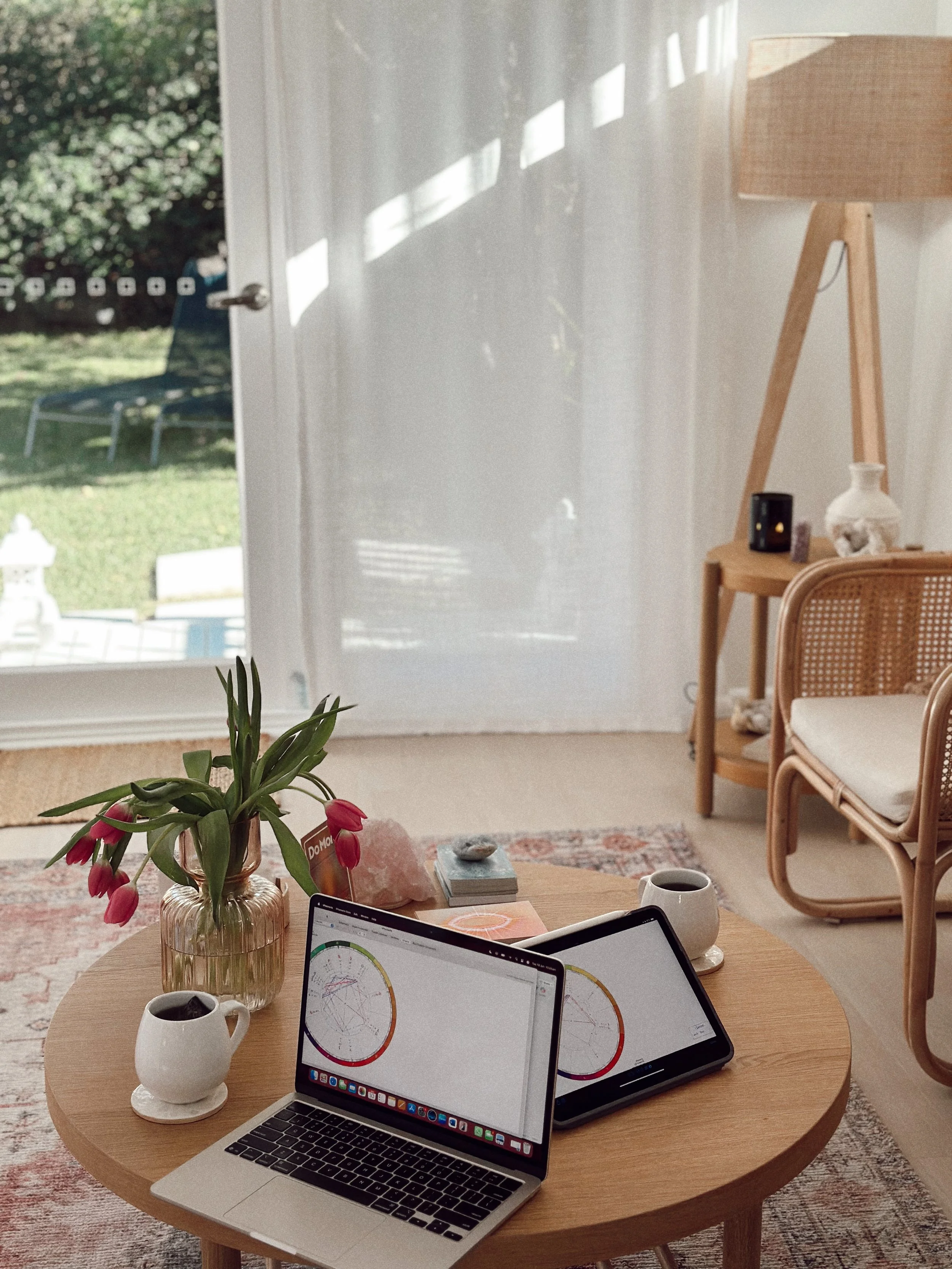 A round wooden table in a bright room with a laptop displaying charts, a tablet, a white mug, a mug of coffee, a vase of pink flowers, and a small rock on it. In the background, there are beige chairs with cane backs and a bookshelf. A large window with white curtains lets in natural light.