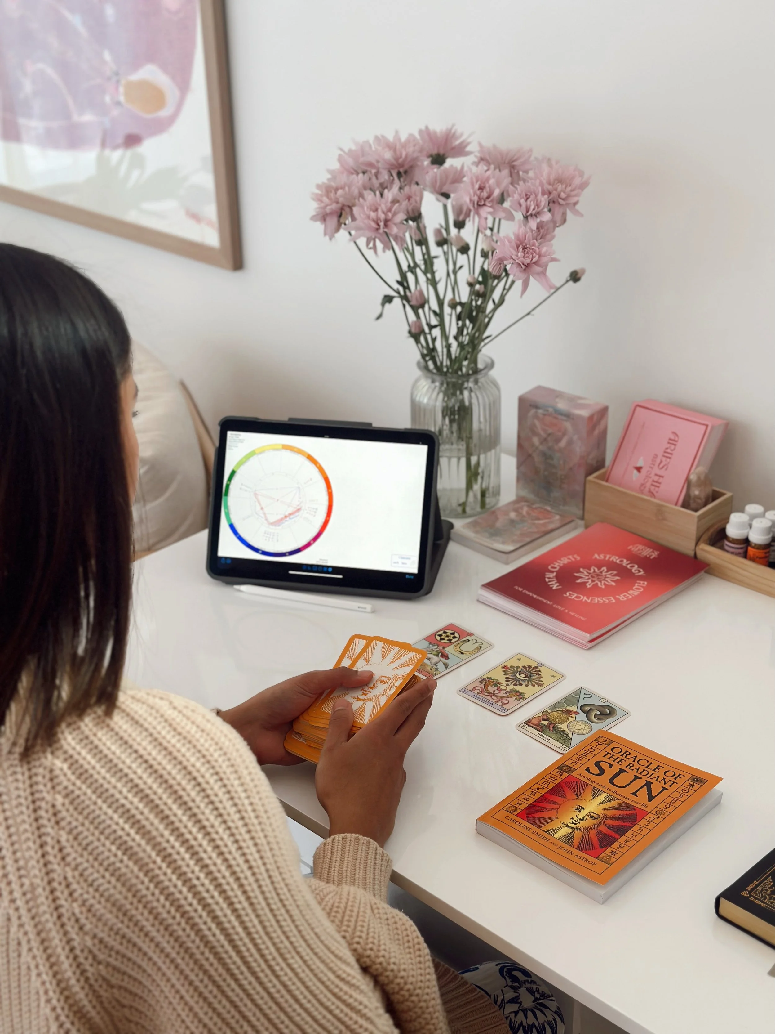 A person playing a card game at a white table, with tarot cards laid out, a tablet displaying a colorful circular chart, and a vase with pink flowers in the background.