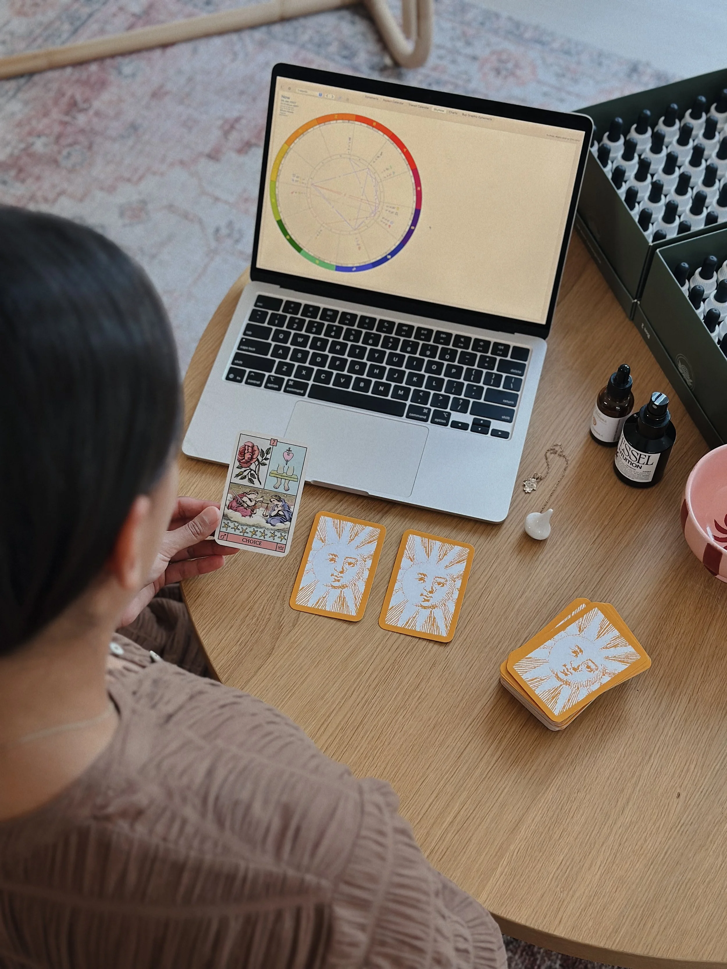 A person sitting at a wooden table, holding tarot cards, with a laptop displaying a colorful chart, bottles of essential oils, and a box of small dropper bottles nearby.