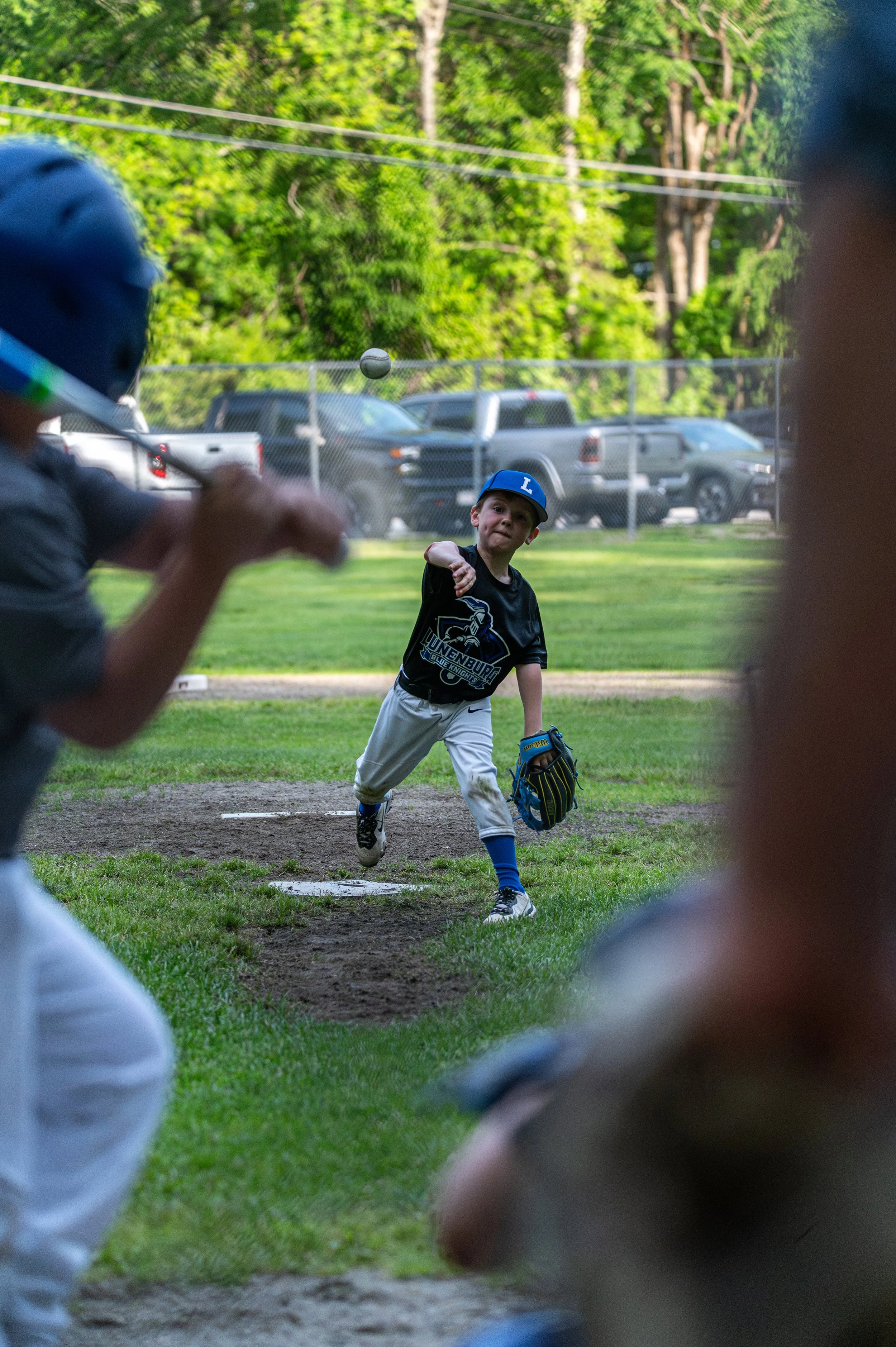 A young boy in a baseball uniform throws a ball during a game, with other players and a fence in the background, surrounded by trees and parked cars.