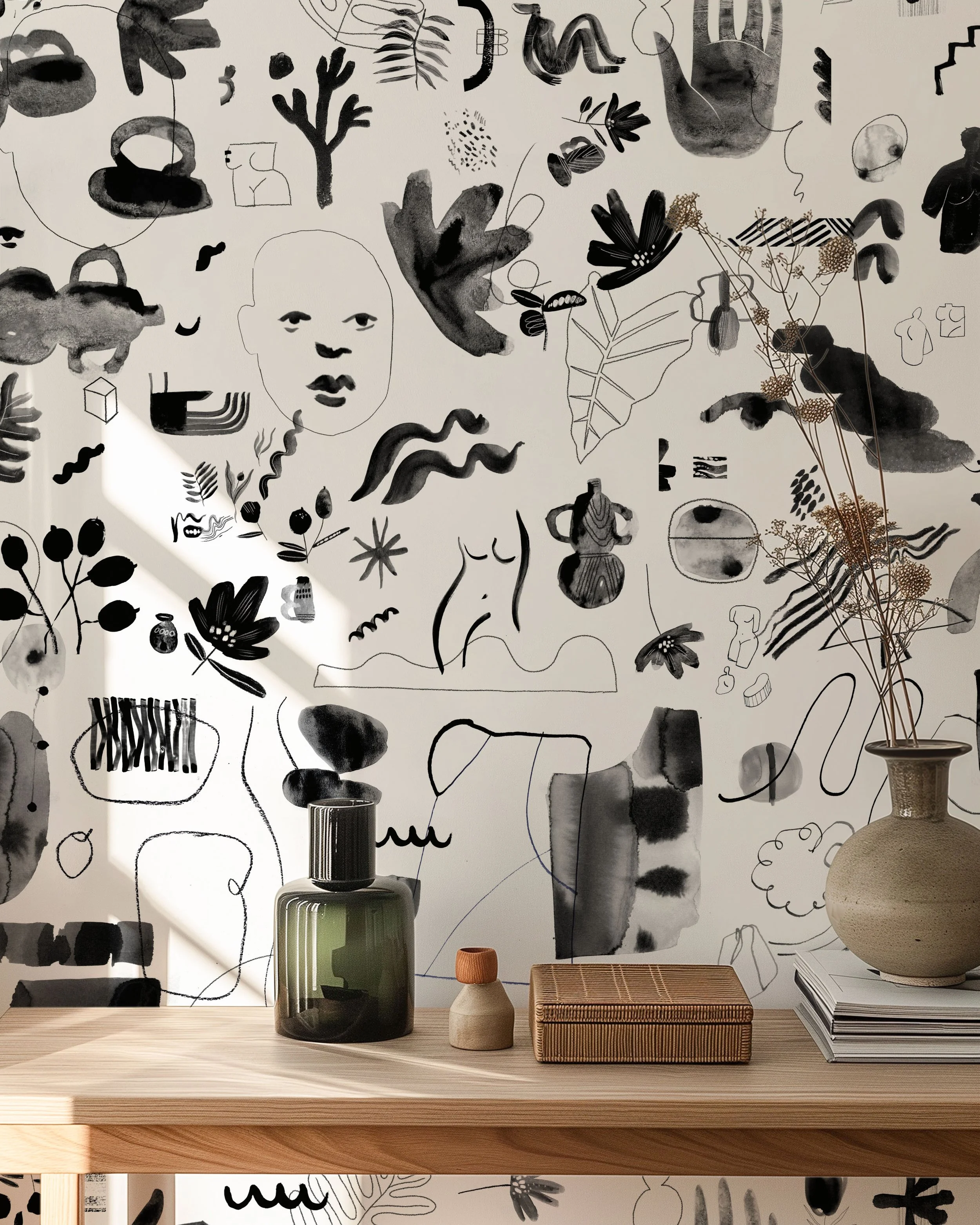 Decorative wall with abstract black and white sketches of faces, plants, and various shapes on a white background. A wooden table in front holds a green glass bottle with a black lid, a small beige and orange ceramic jar, a woven box, and a beige vase with dried foliage.