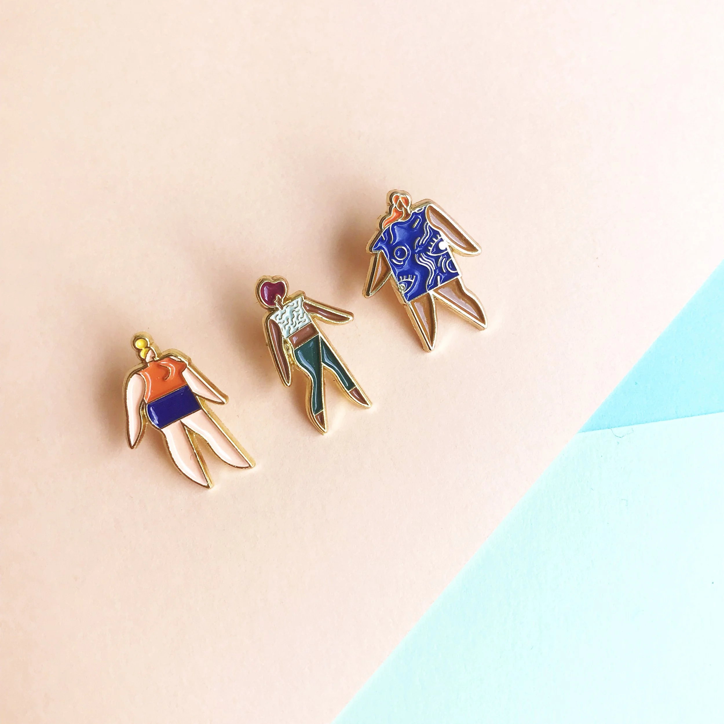 Three colorful enamel pins of abstract human figures on pastel pink and blue background.