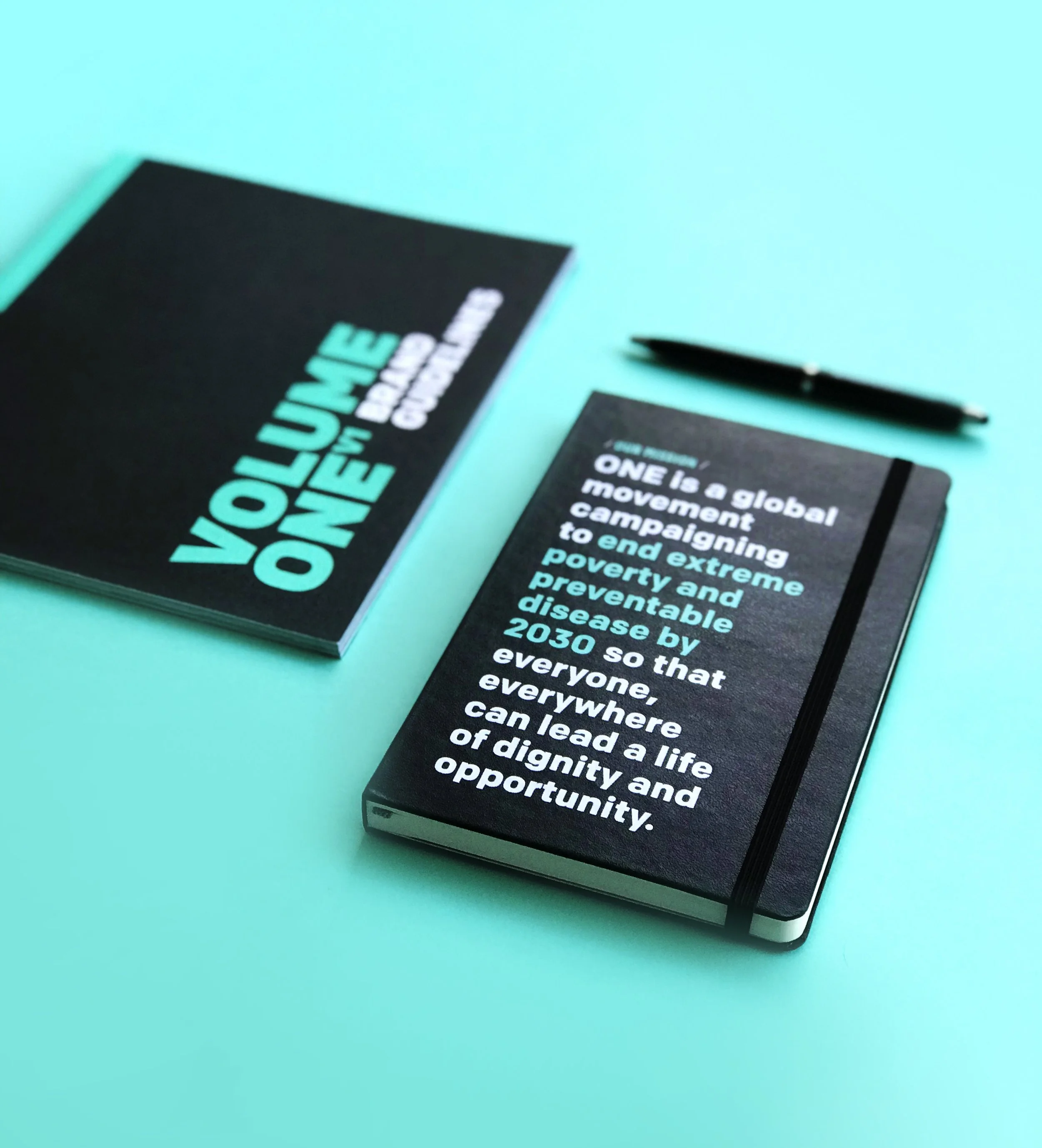Two black notebooks with motivational messages, one with the text 'VOLUME ONE' and a blue pen on a light blue surface.