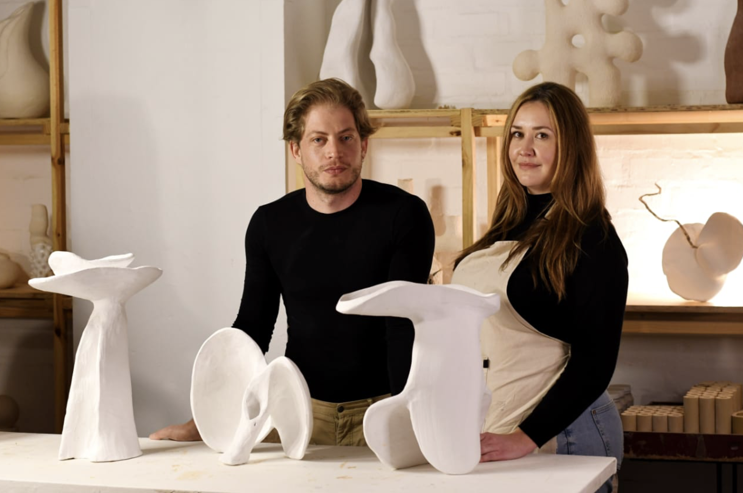 A man and woman in a pottery studio, standing behind a table with ceramic sculptures and pottery pieces.