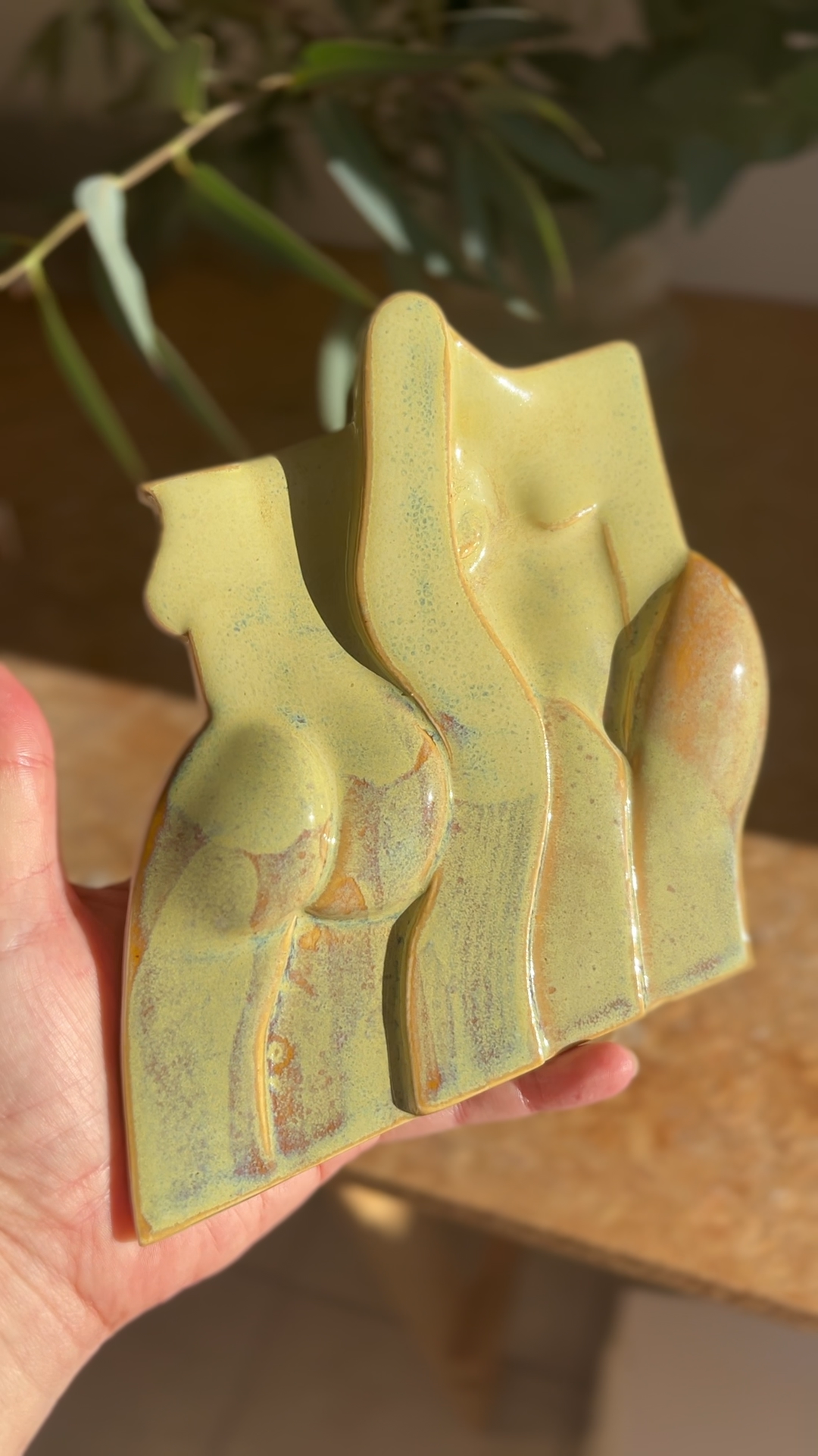A green abstract ceramic sculpture resembling a woman with rounded breasts and a curvy form, held in a person's hand, with a blurred background of leaves and a table.