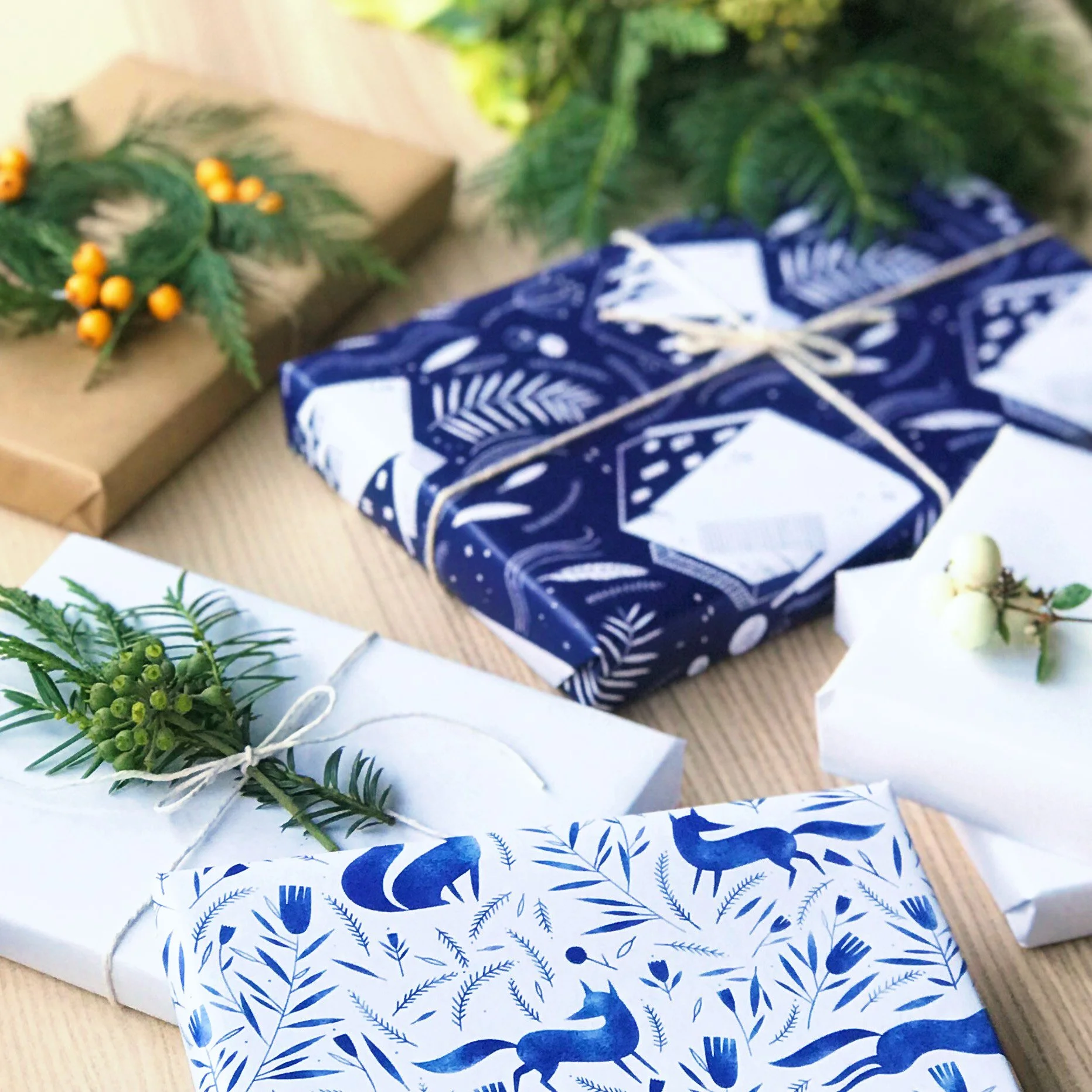 Christmas wrapped gifts with white, blue, and brown paper, decorated with sprigs of greenery and berries, on a wooden surface.